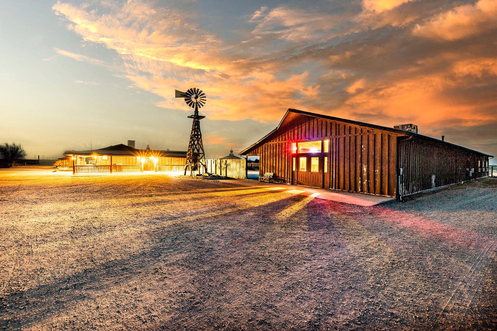 Sunset over a rural farmyard with a metal windmill and wooden buildings.