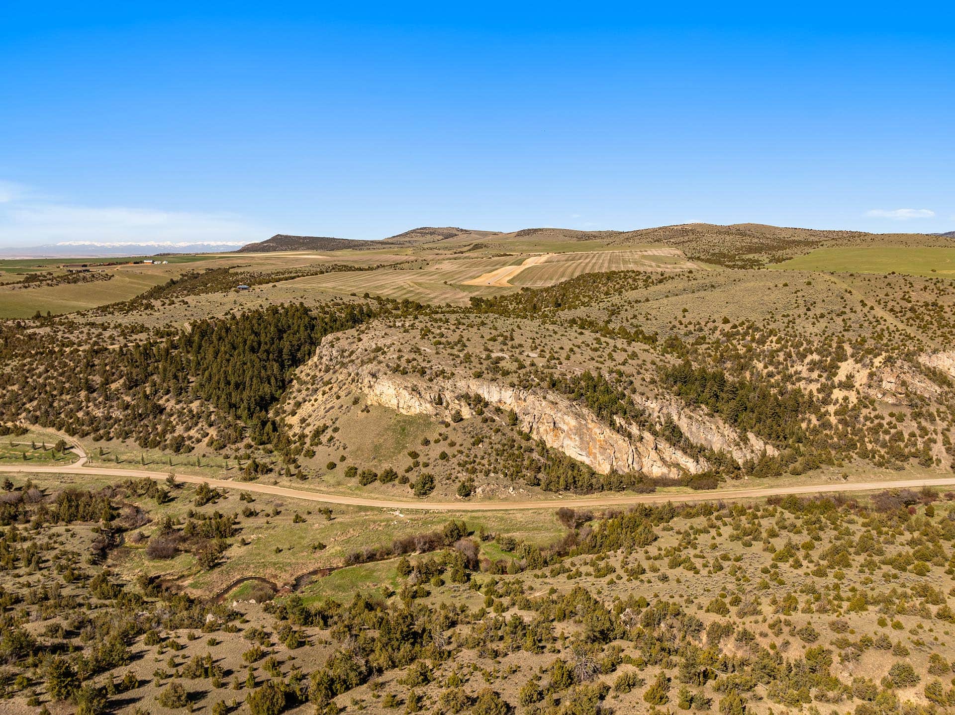 Wide rural landscape with rolling fields, scattered trees, and a winding road under a bright blue sky.