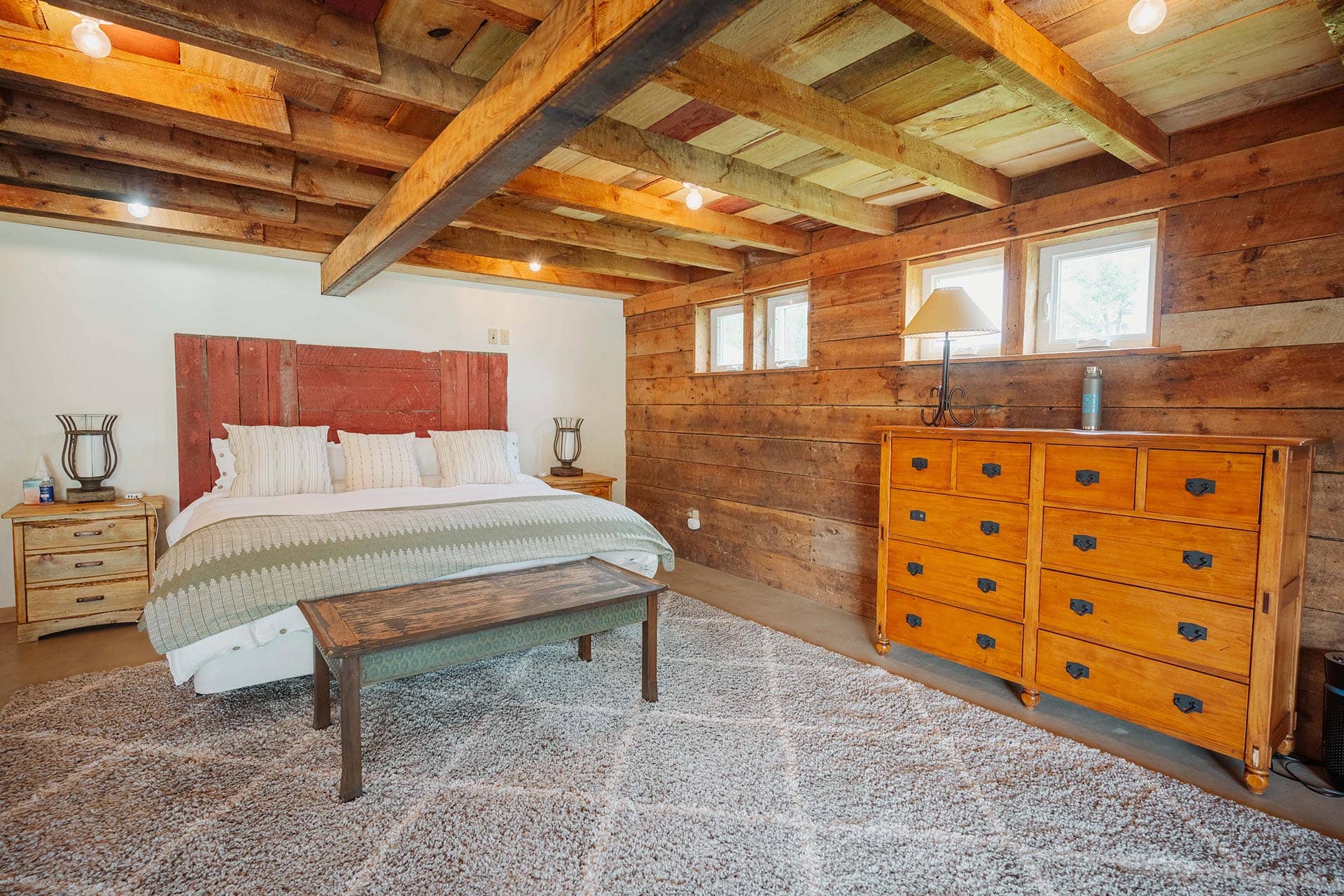 Rustic bedroom with exposed wooden beams, a red headboard, white bedding, and a large wooden dresser.