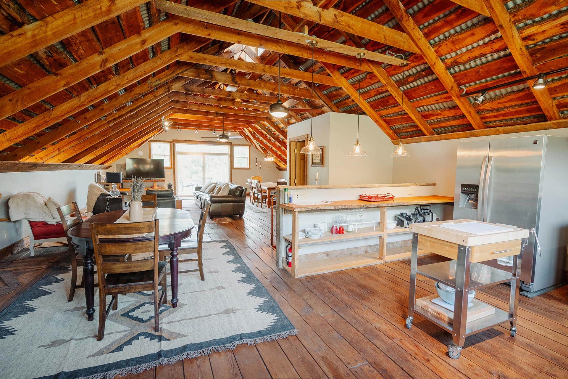 Open-concept loft with exposed wooden beams, a kitchen island, dining table, and seating area in a rustic home.