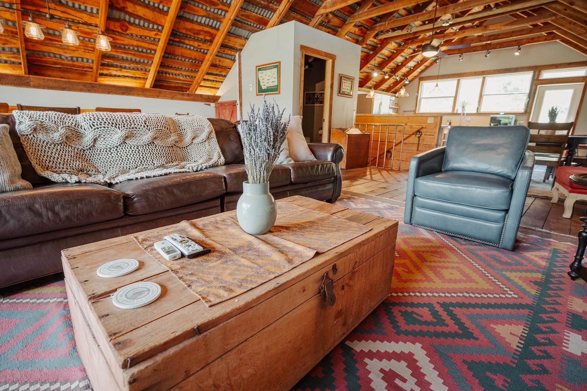 Cozy loft living room with exposed wooden beams, brown leather sofa, chunky knit blanket, and a rustic wooden coffee table on a colorful rug