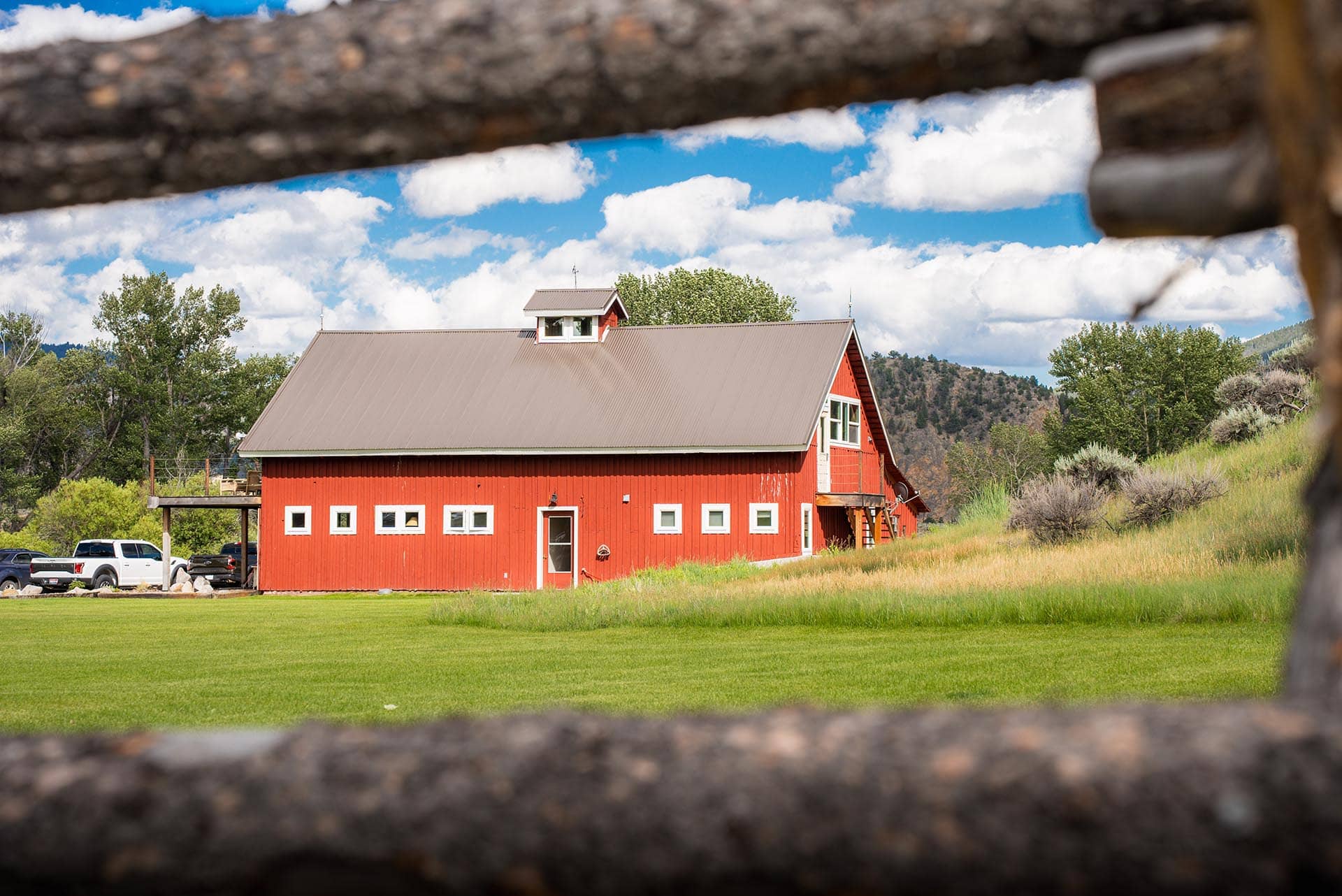 Red barn with gray metal roof in a green field, under a blue sky with fluffy white clouds, framed by a blurred foreground log.