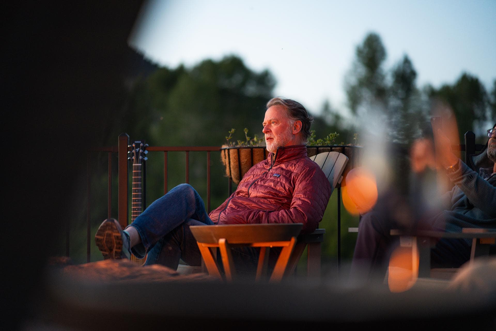 Older man in a red jacket relaxing on an outdoor deck with a guitar nearby and trees in the background.