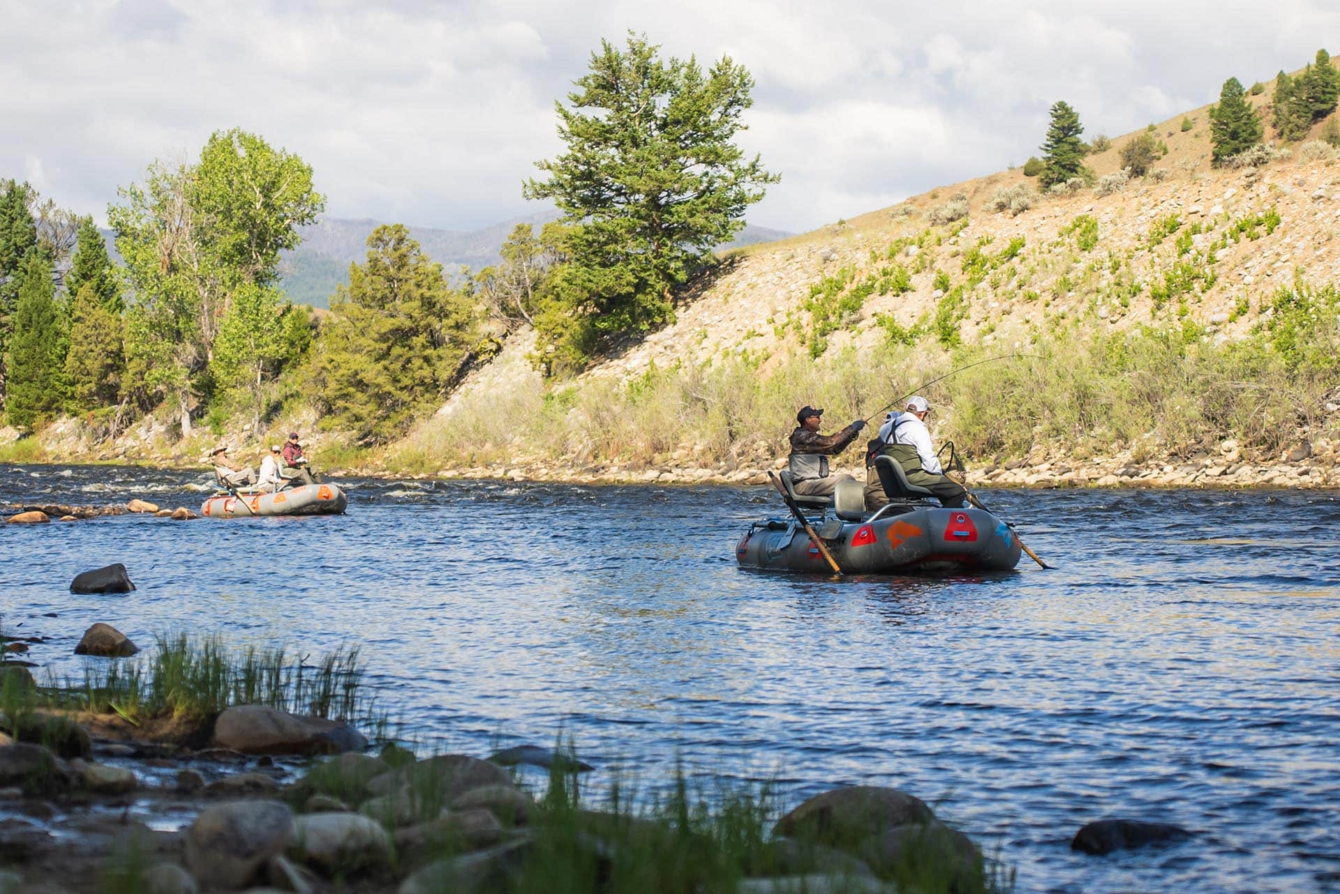Two inflatable dinghies carrying four people fishing on a wide river, wearing life jackets, with rocky hillside and trees in the background.