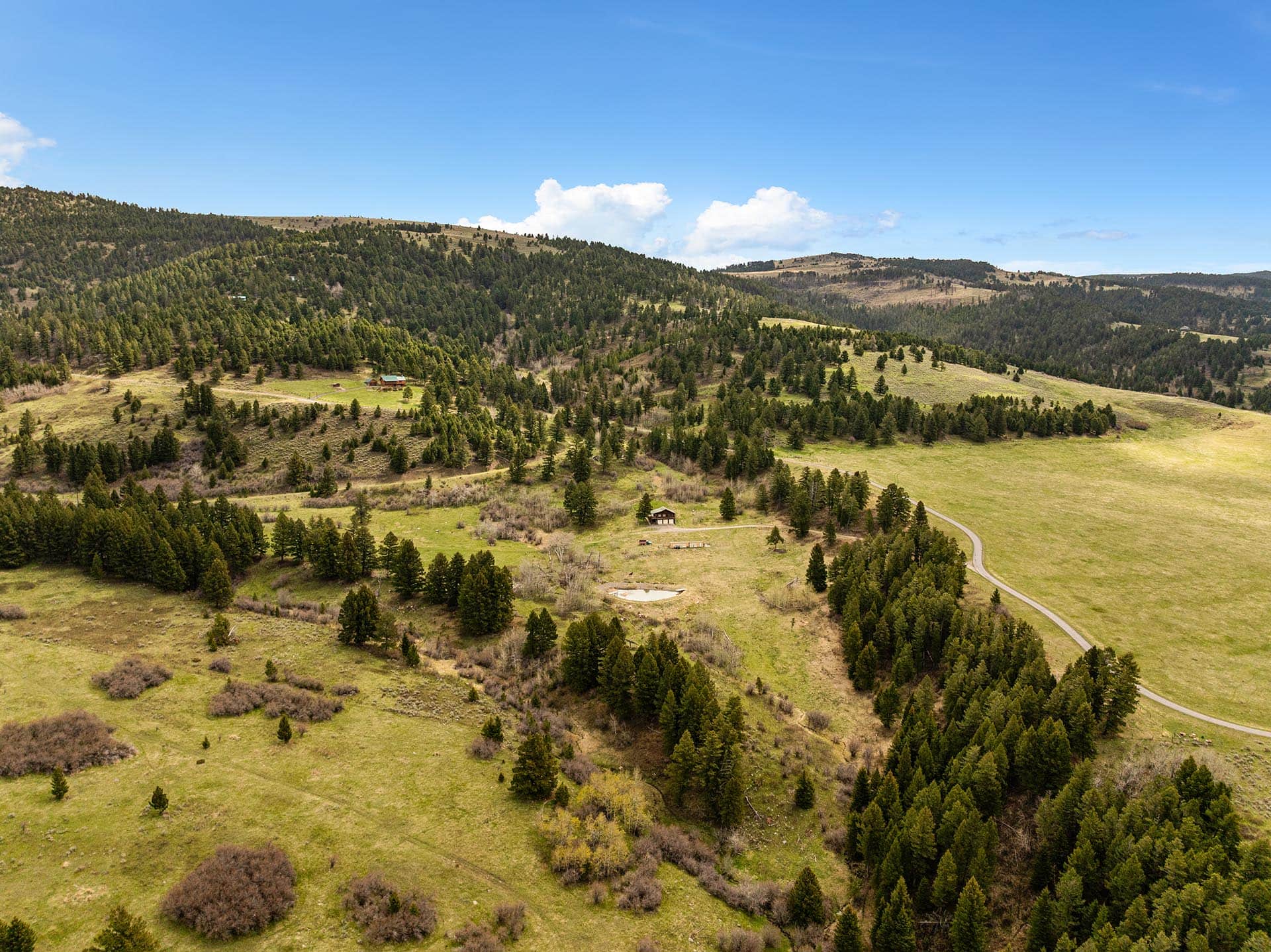 Aerial view of rolling hills with conifer forests, scattered houses, and a winding road under a clear blue sky at midday.