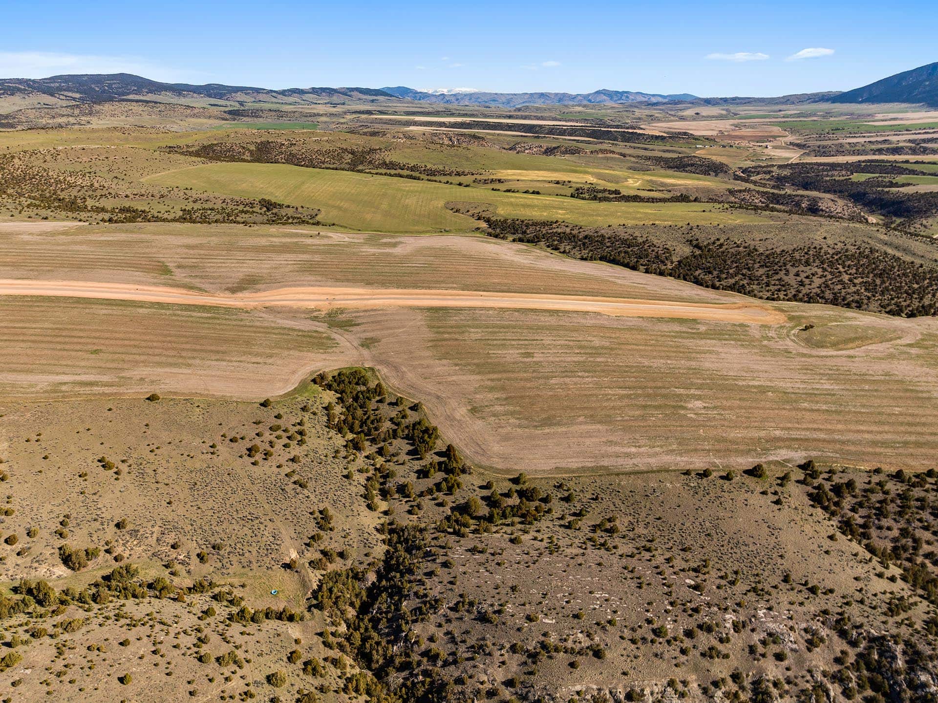 Arid rolling countryside with patchy fields, dirt roads, and scattered shrubs under a clear blue sky.