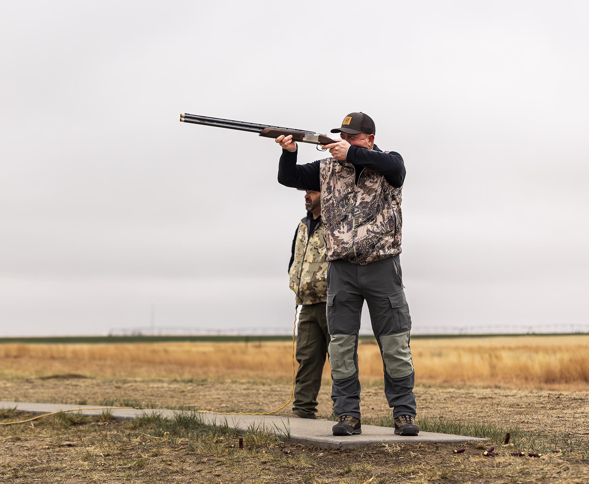 Man in a camouflage vest and cap aiming a double-barreled shotgun while standing on a grassy field, with another man in the background watching nearby.