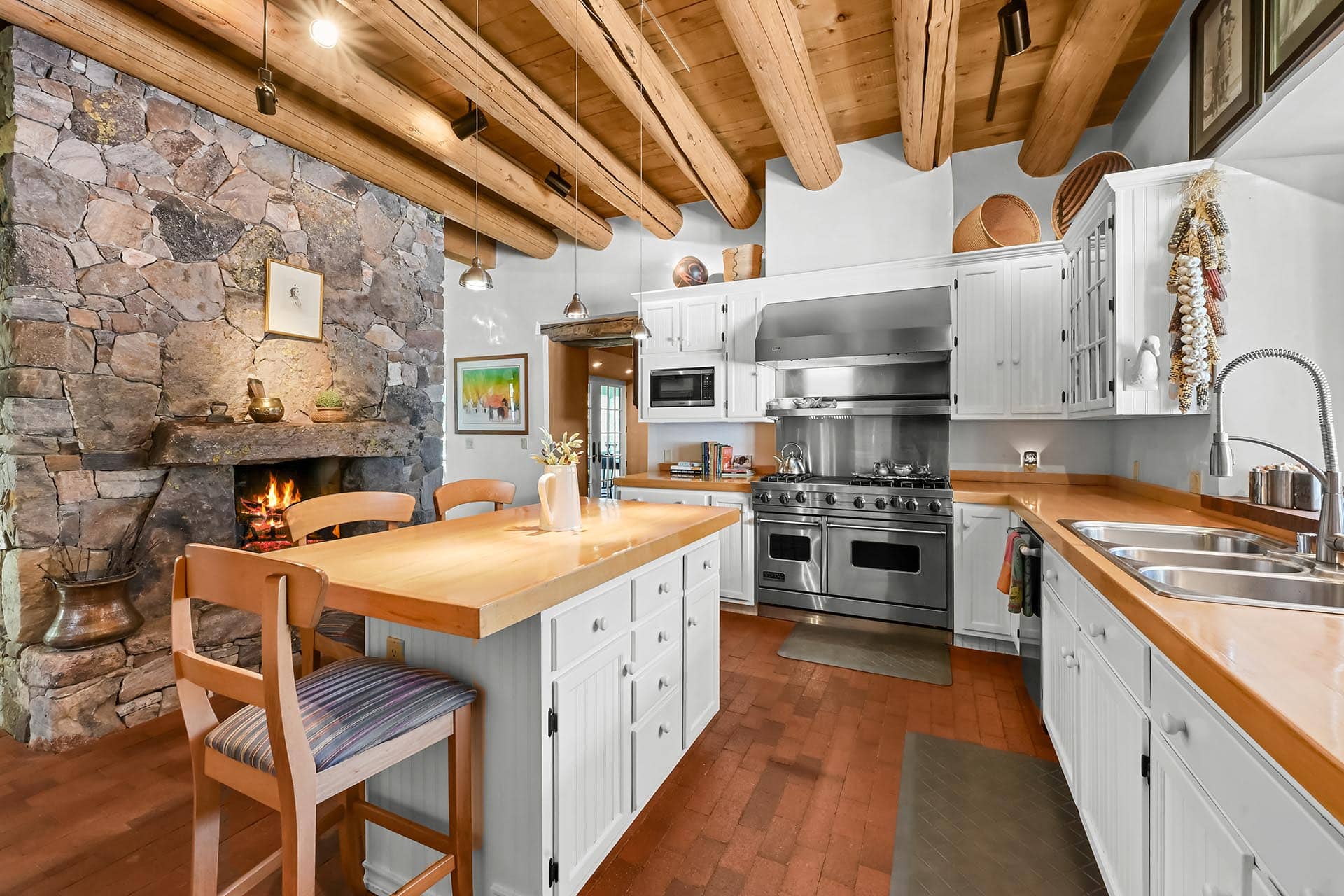 Cozy rustic kitchen with a stone fireplace, wooden beams, white cabinetry, and stainless steel appliances.