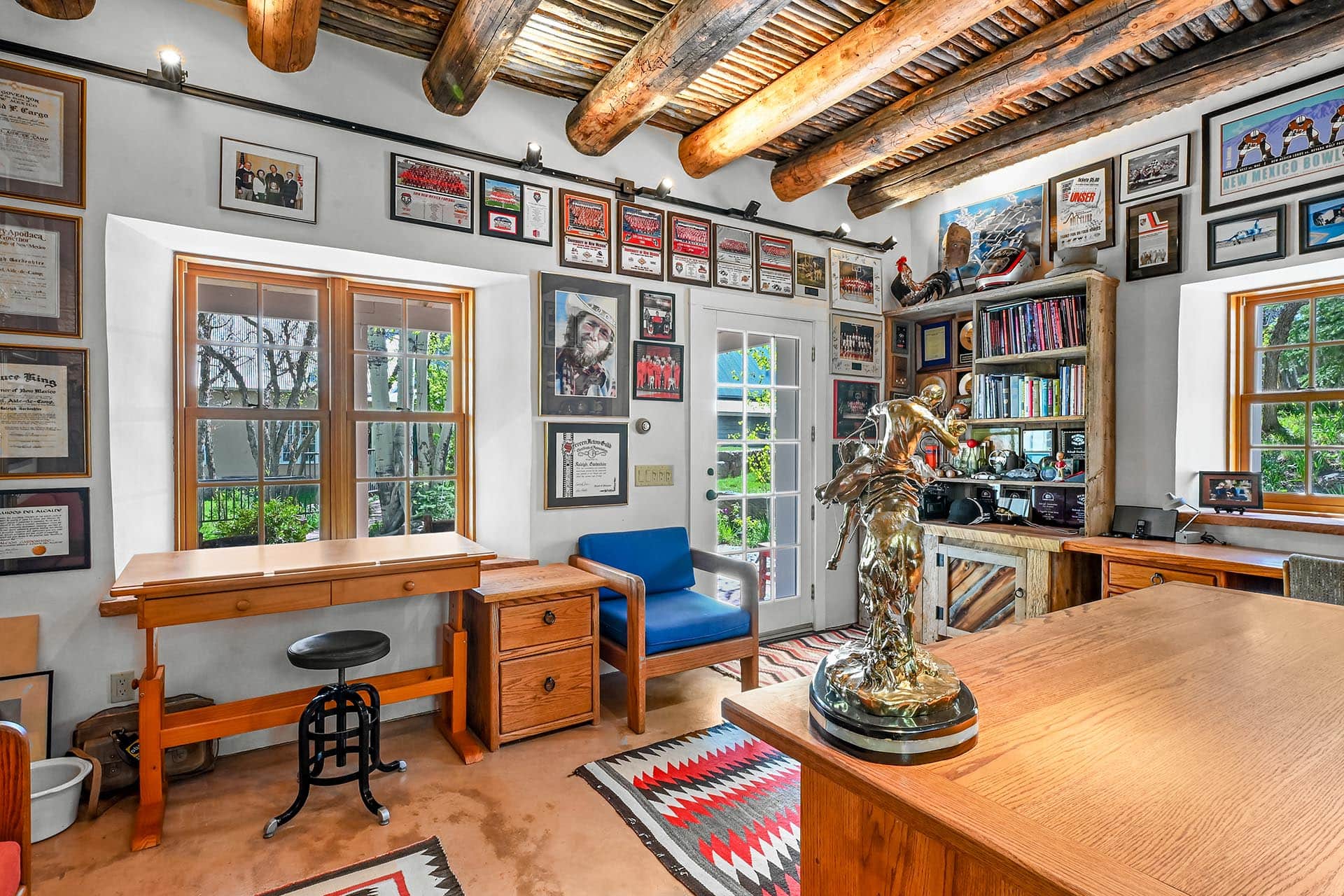 Home office with wood beams, wall of framed awards and photos, wooden desks, and a metallic statue on the foreground table.