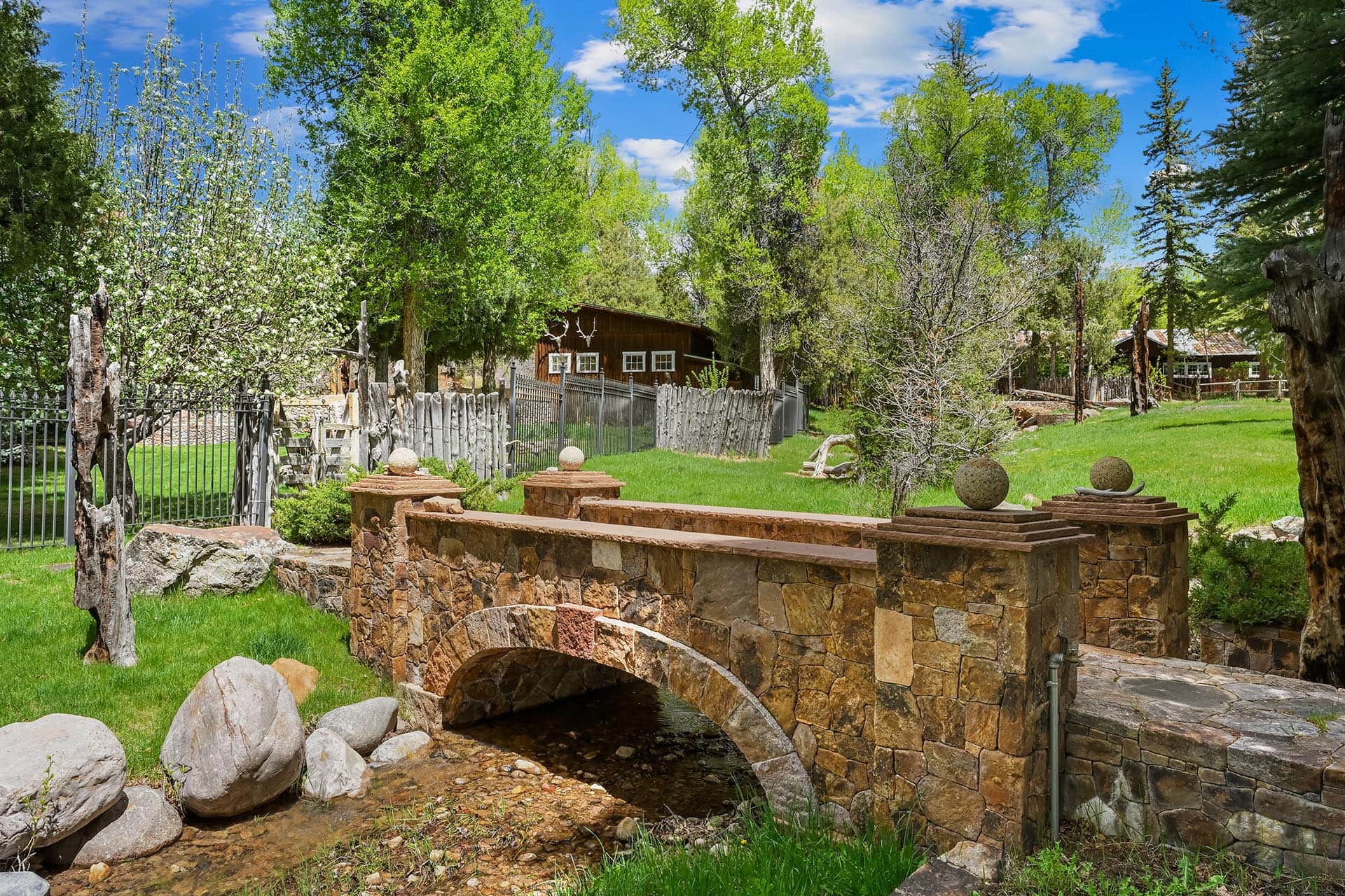 Stone arch bridge over a shallow creek in a lush green garden with trees and a wooden fence in the background