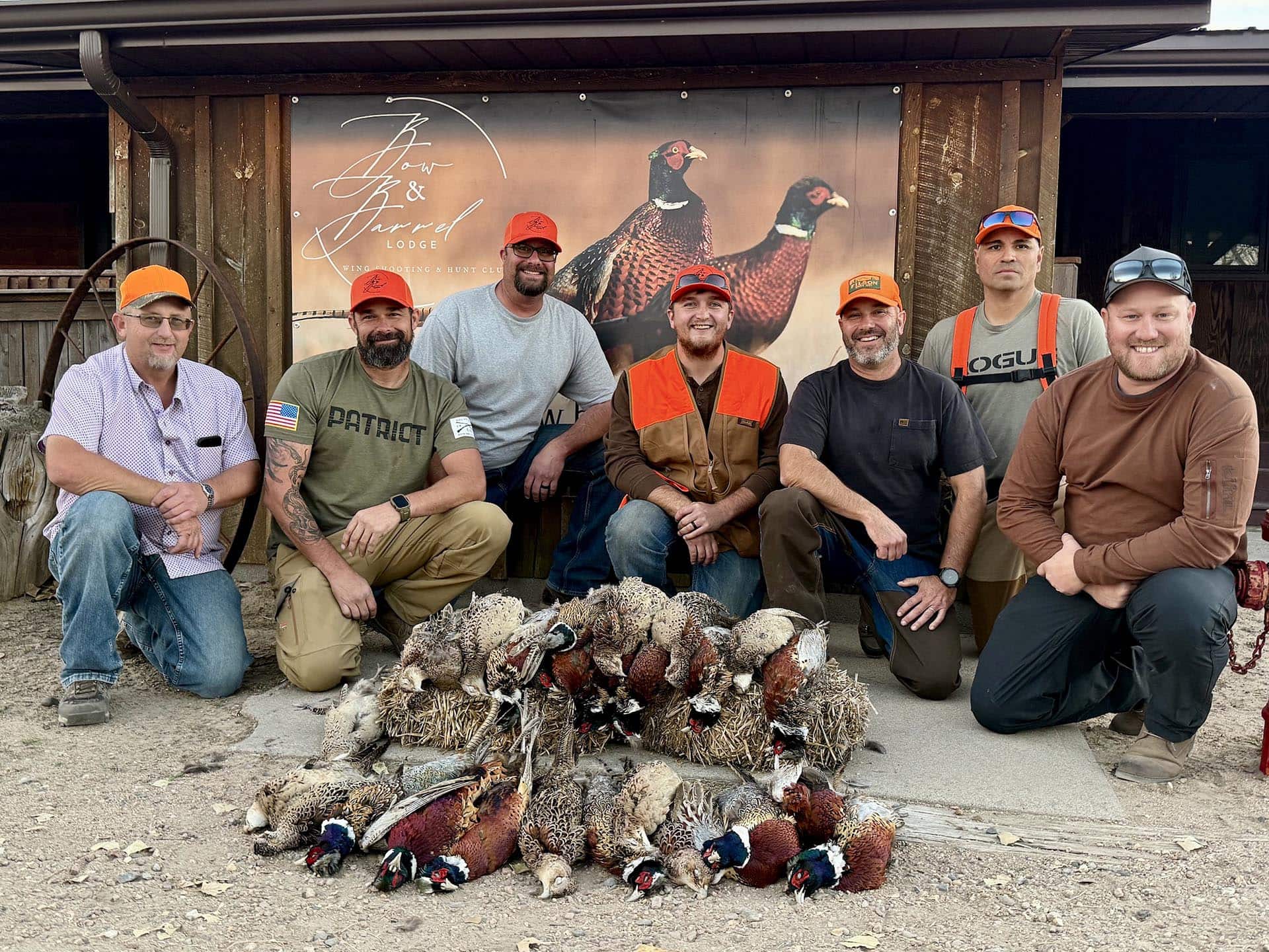 Group of six men in hunting gear posing with a row of pheasants on the ground outside a lodge sign.