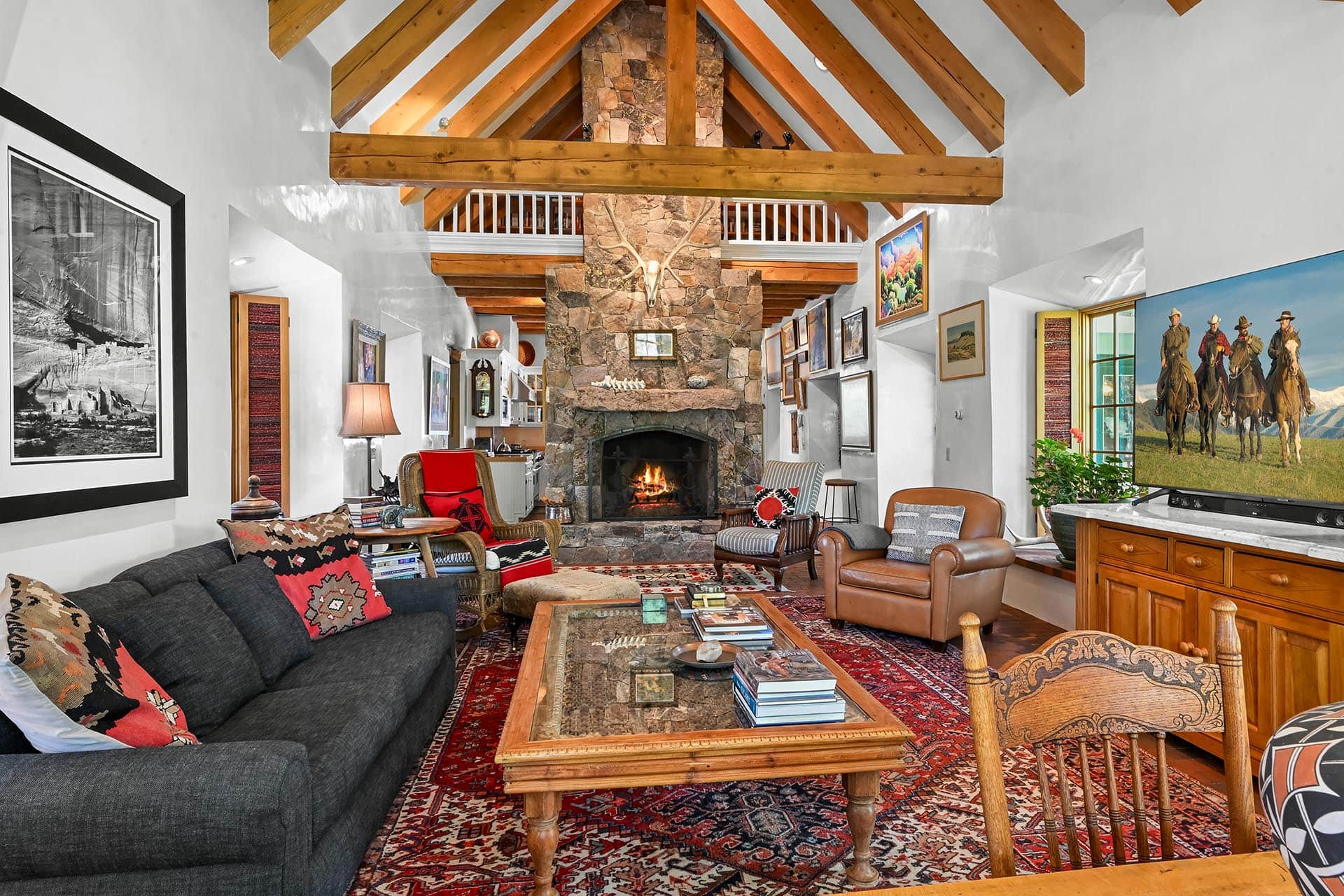 Cozy living room with a stone fireplace, exposed wooden beams, and a large TV showing riders on horseback.