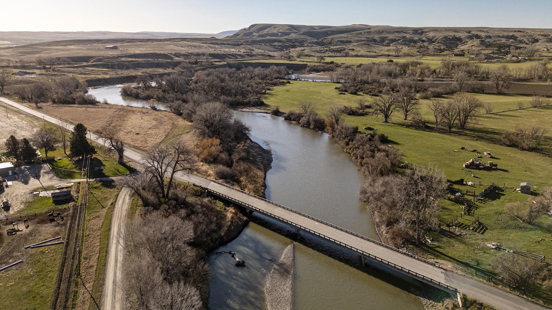 Aerial view of a winding river with a bridge crossing a rural valley, fields and leafless trees under a clear sky.