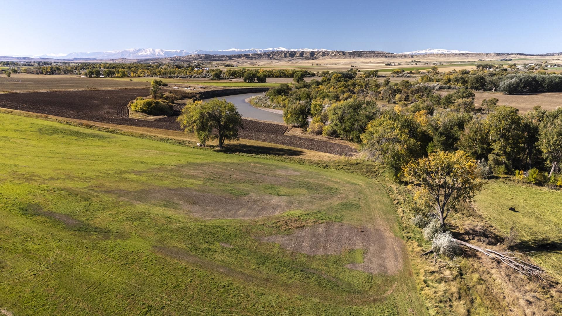 Aerial view of a winding river through farmland, with fields, trees, and distant snow‑capped mountains under a clear blue sky.