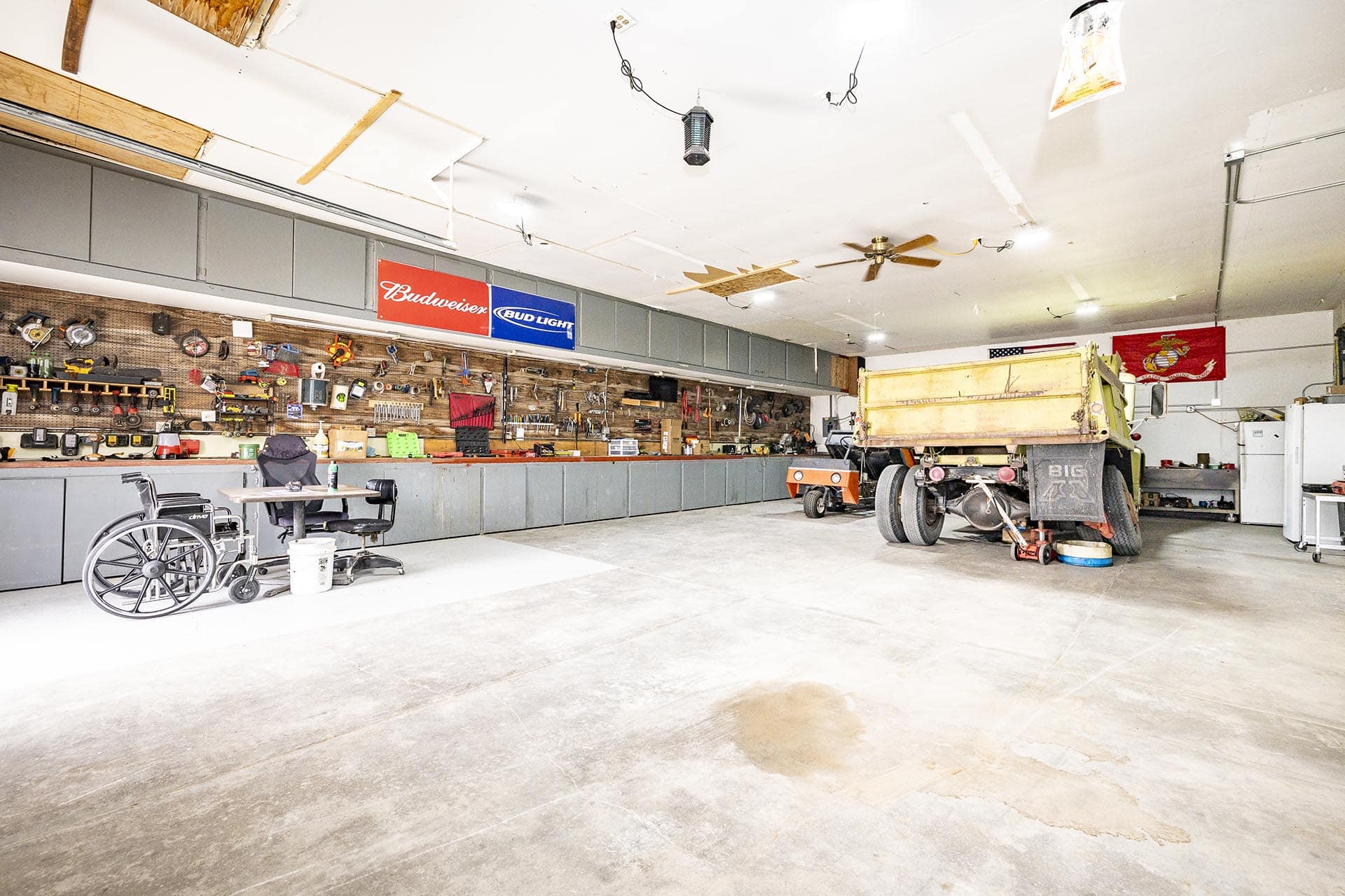 Spacious workshop with a long tool wall, workbench, and a large dump truck trailer inside; wheelchair and office chair in the foreground.