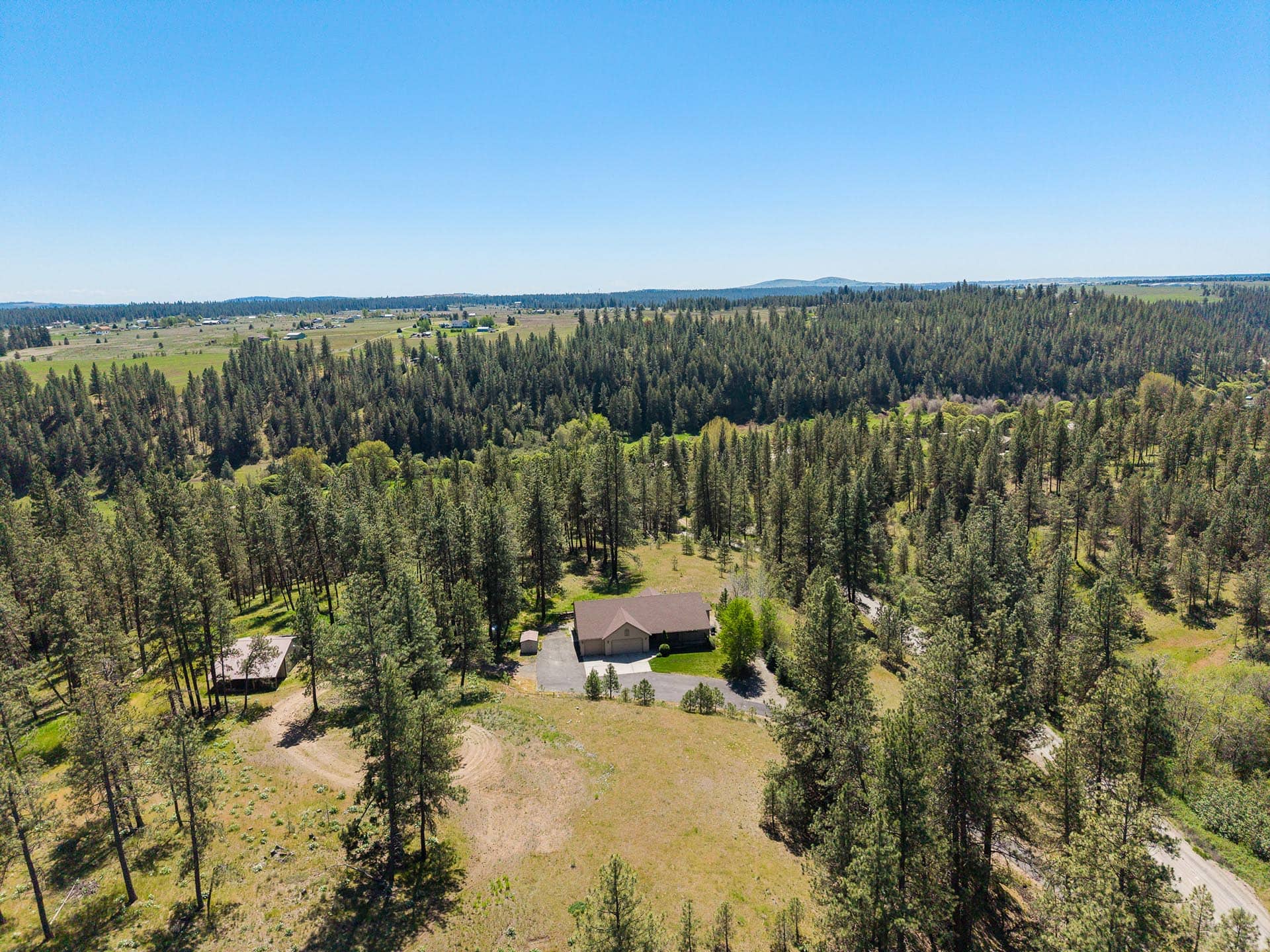 Aerial view of a forested rural neighborhood with scattered houses and open fields under a clear blue sky.