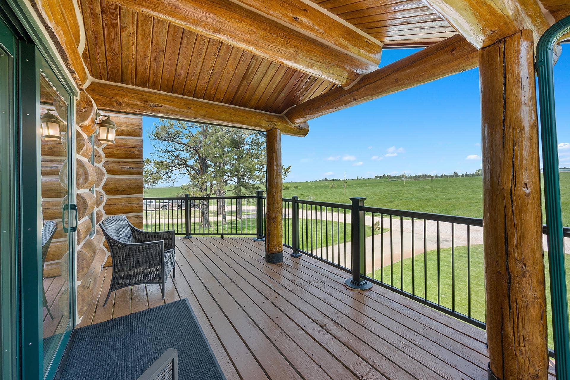 Wooden log cabin porch with seating and a railing, overlooking a grassy field under a blue sky.