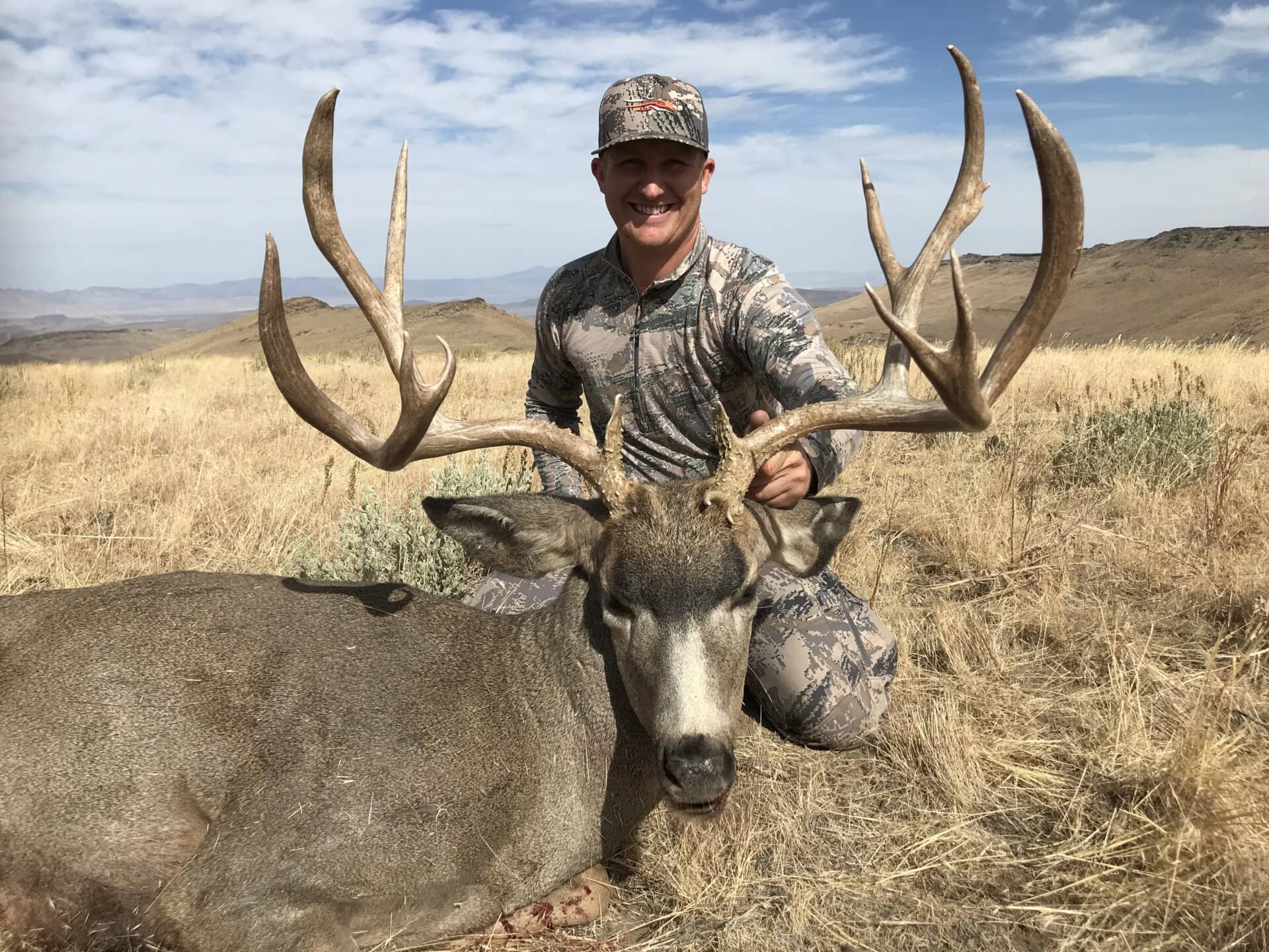 Hunter in camo posing with a large antlered deer in a dry grassy field under a blue sky—outdoors landscape image