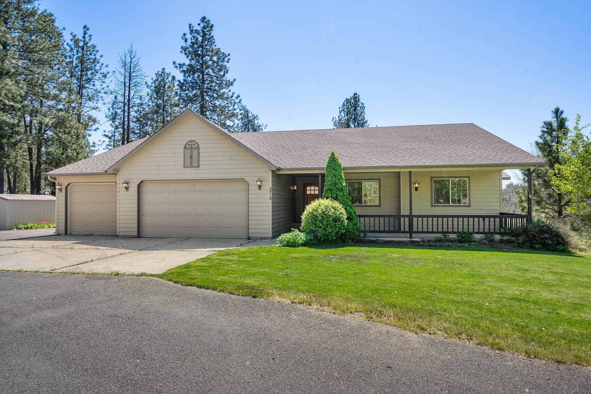 Single-story beige house with a two-car garage, front porch, and well-kept lawn on a sunny day, with tall trees in the background.