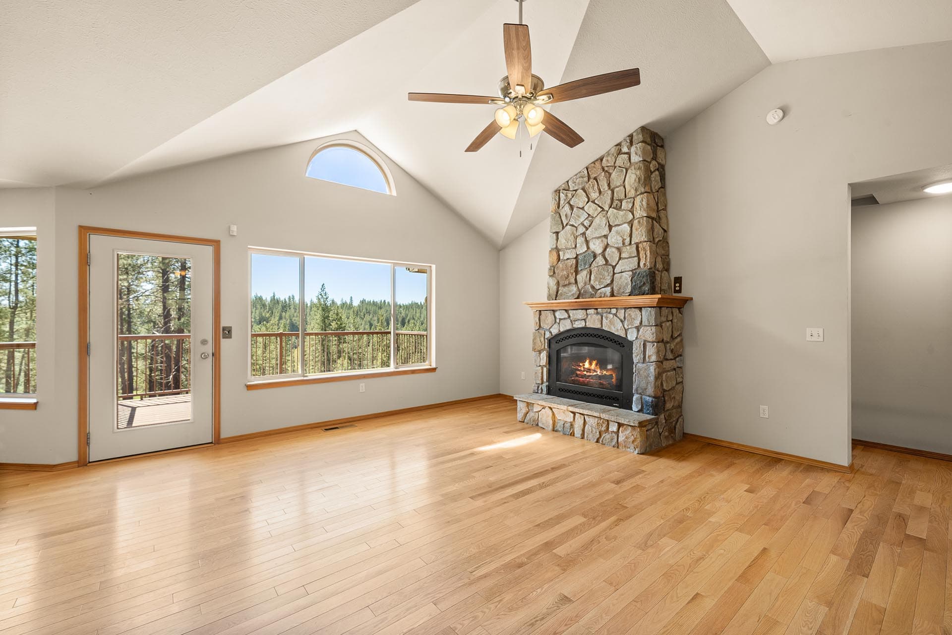 Open living room with vaulted ceiling, stone fireplace, and hardwood floors; door to a deck and forest view.