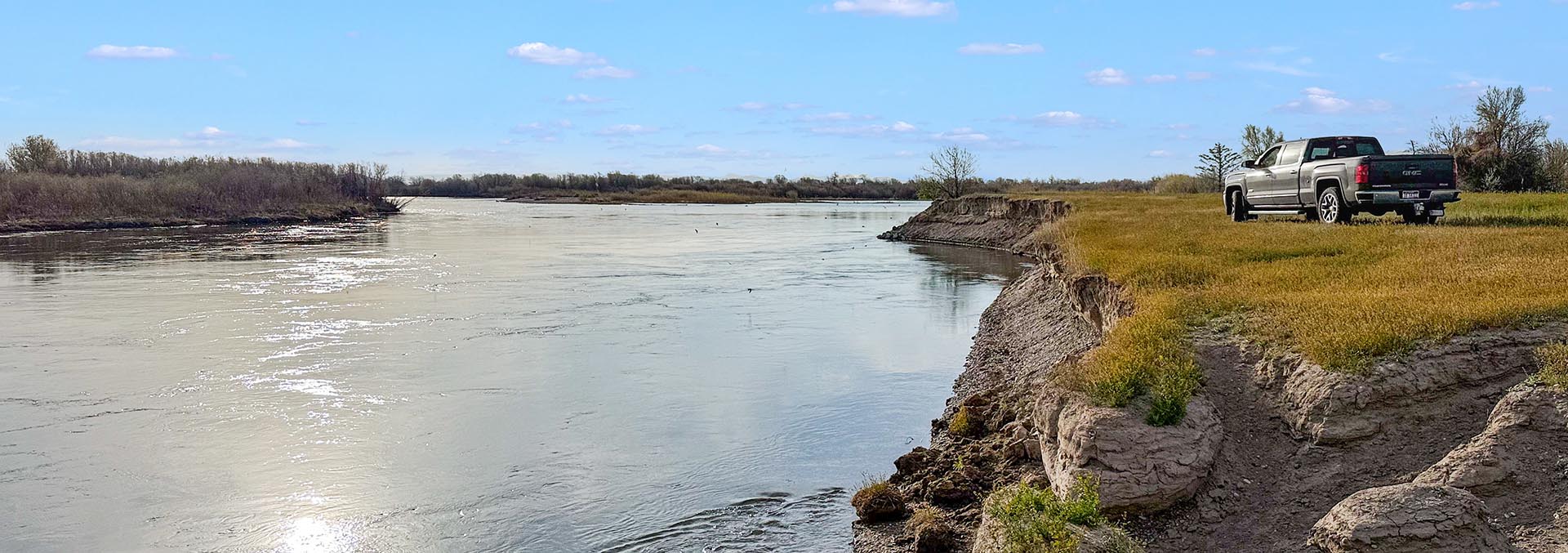 Silver pickup truck parked on a grassy riverbank, overlooking a wide calm river under a blue sky with scattered clouds.