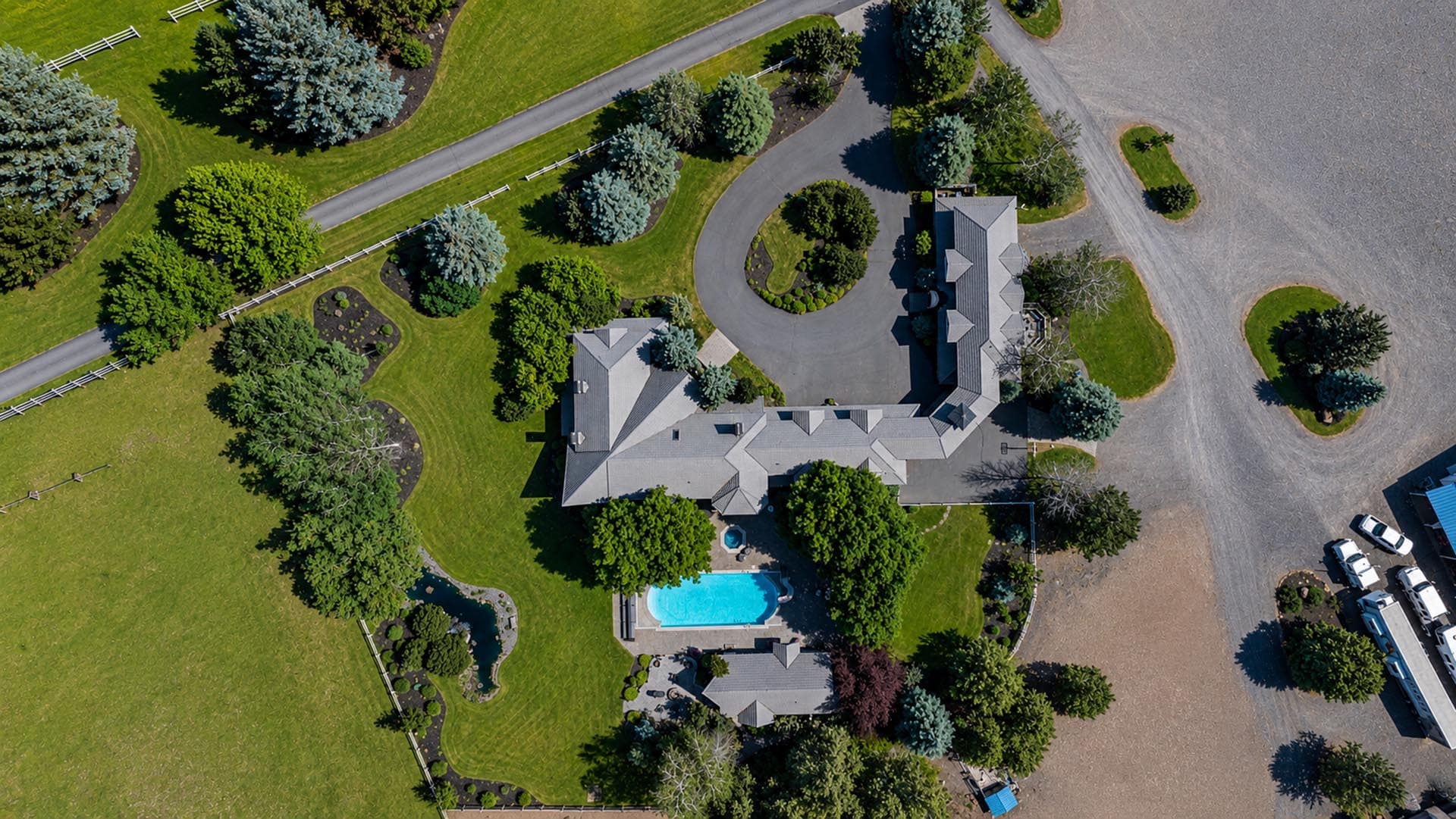 Aerial view of a large estate with a gray-roofed house, curved circular driveway, and surrounding green lawns and trees, plus a blue swimming pool.