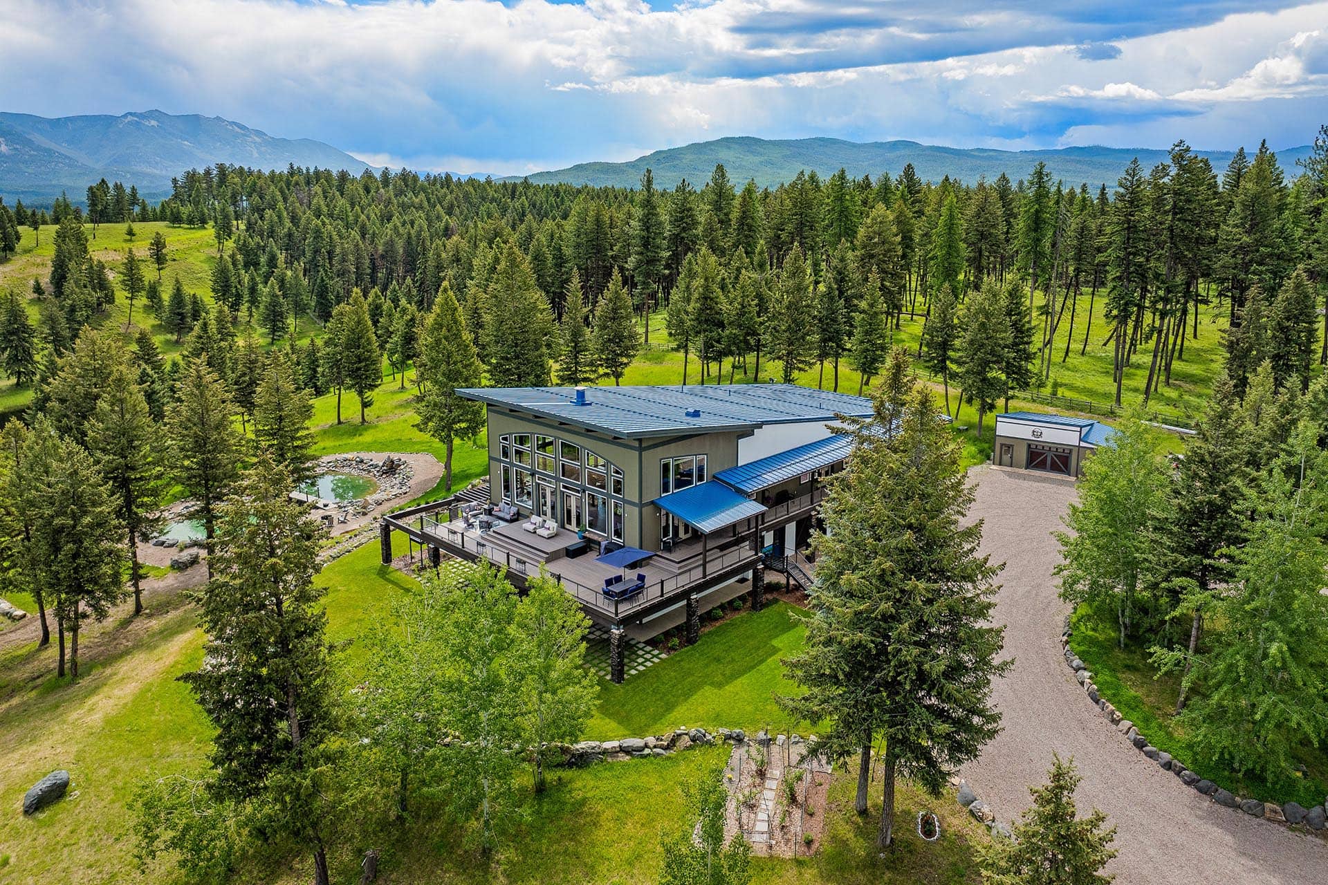 Modern two‑story house with large glass walls and a blue metal roof, set among pine trees with mountains in the distance.