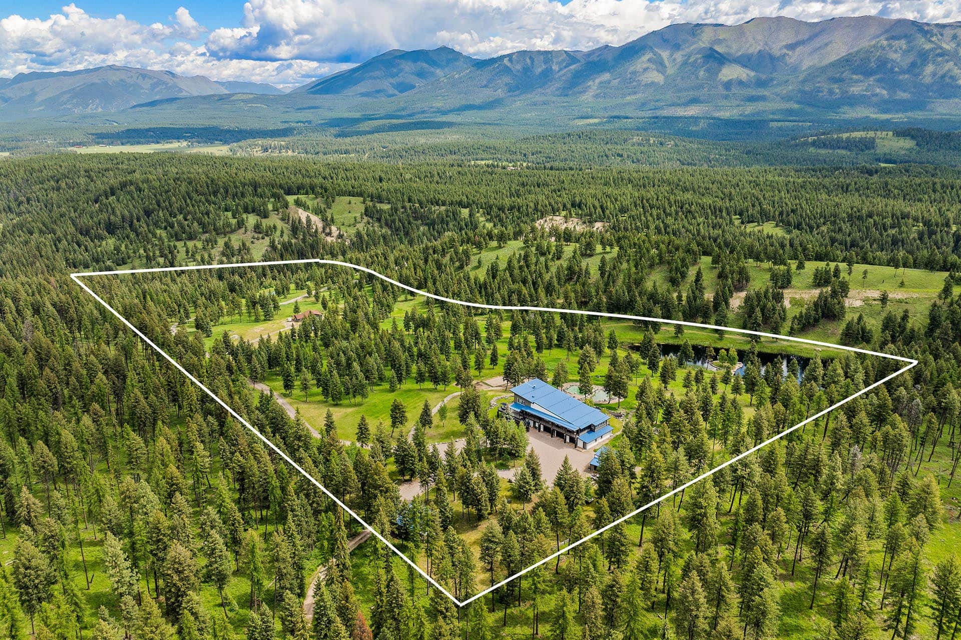 Aerial view of a forested area with a light-colored property boundary outline surrounding a building with a blue roof and surrounding trees, mountains in the distance.