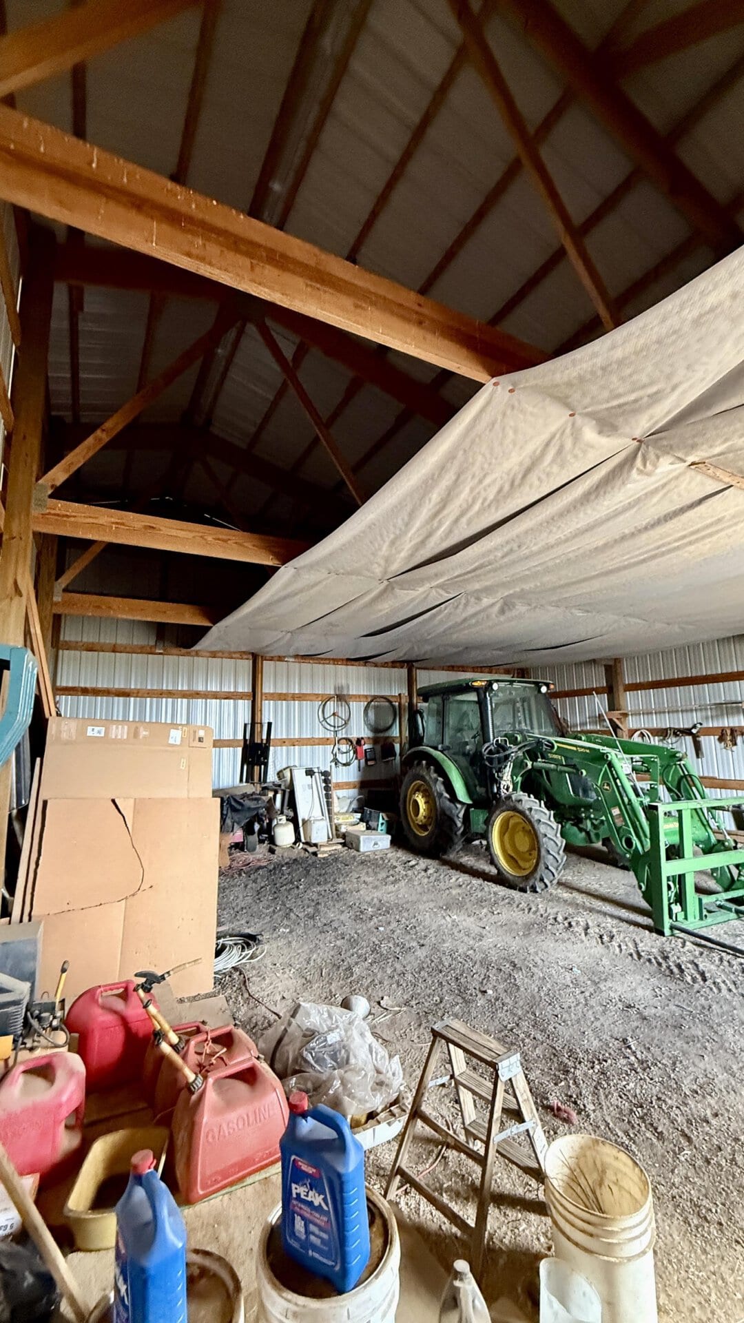 Inside a barn with a green tractor, wooden beams, and a suspended white tarp overhead, cluttered with equipment and boxes.