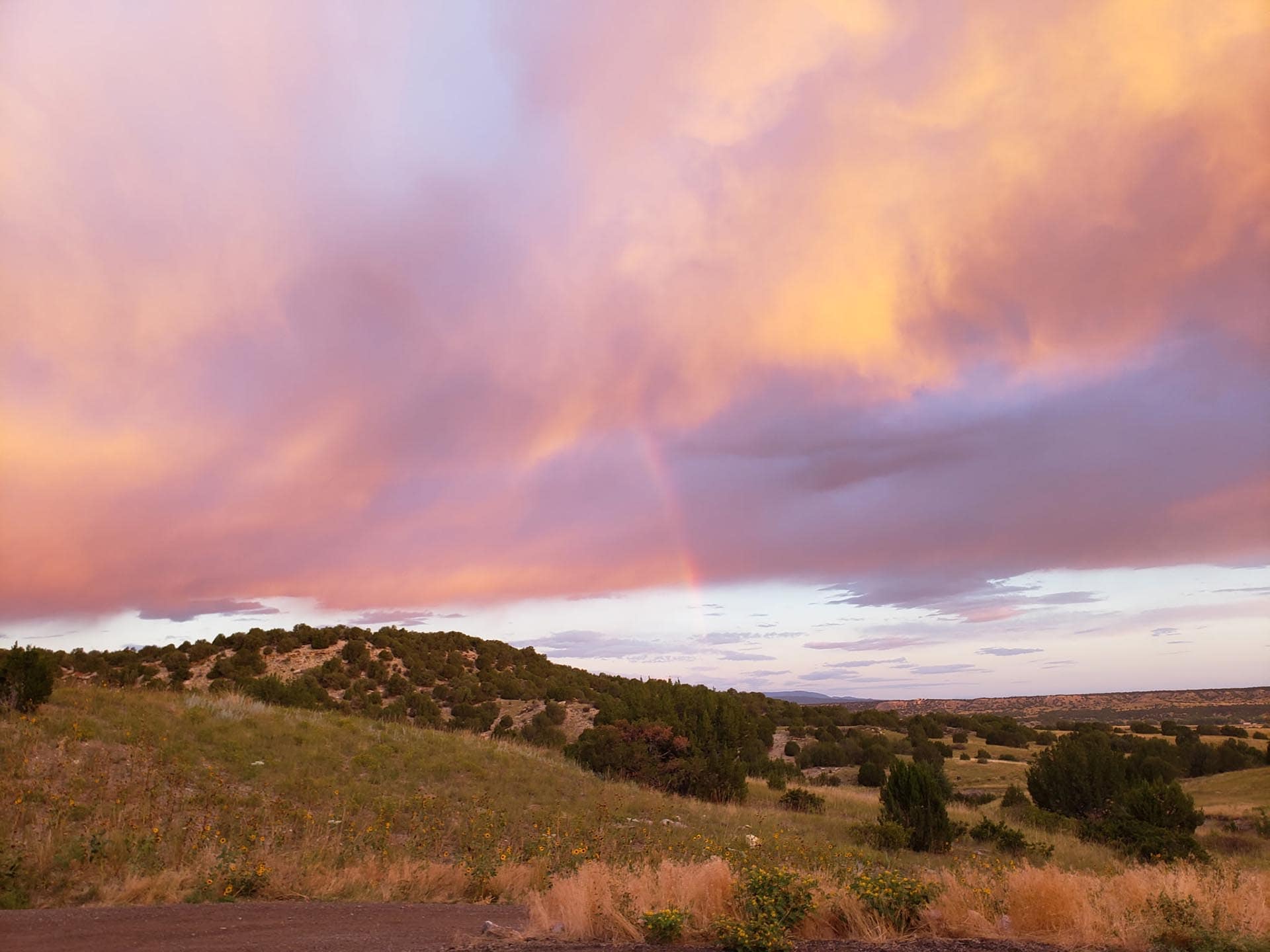 Pastel sunset sky with pink and orange clouds over rolling hills and a faint rainbow above the landscape.
