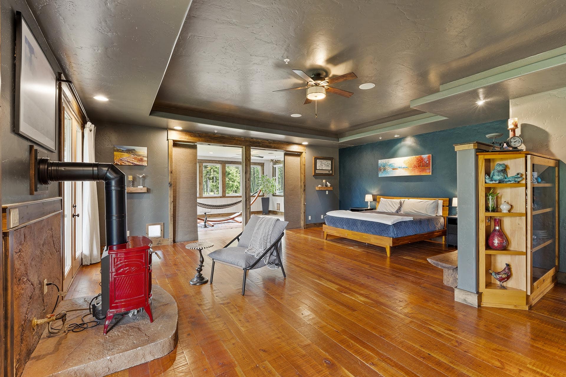 Cozy rustic bedroom with hardwood floors, a red wood stove in the foreground, and a bed against a blue wall; doorway reveals a sunlit sitting area outside.