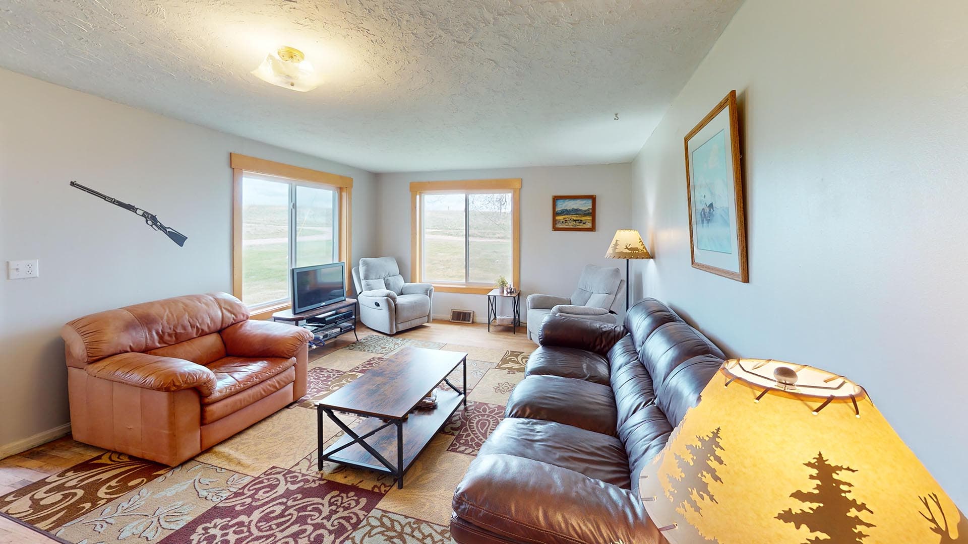 Cozy living room with peach leather sofa, dark brown leather sofa, coffee table, TV stand, and large windows letting in light. A rifle decal is on the wall beside the outlet.