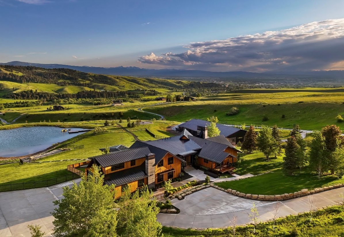 Lodge-style wooden house cluster on a green hillside with a pond, trees, and rolling fields.