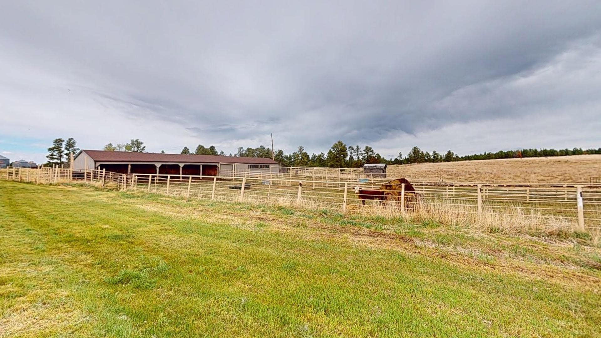 Rural farm scene with a long barn, wooden fences, and a horse in a pen on a grassy field under gray, cloudy skies.