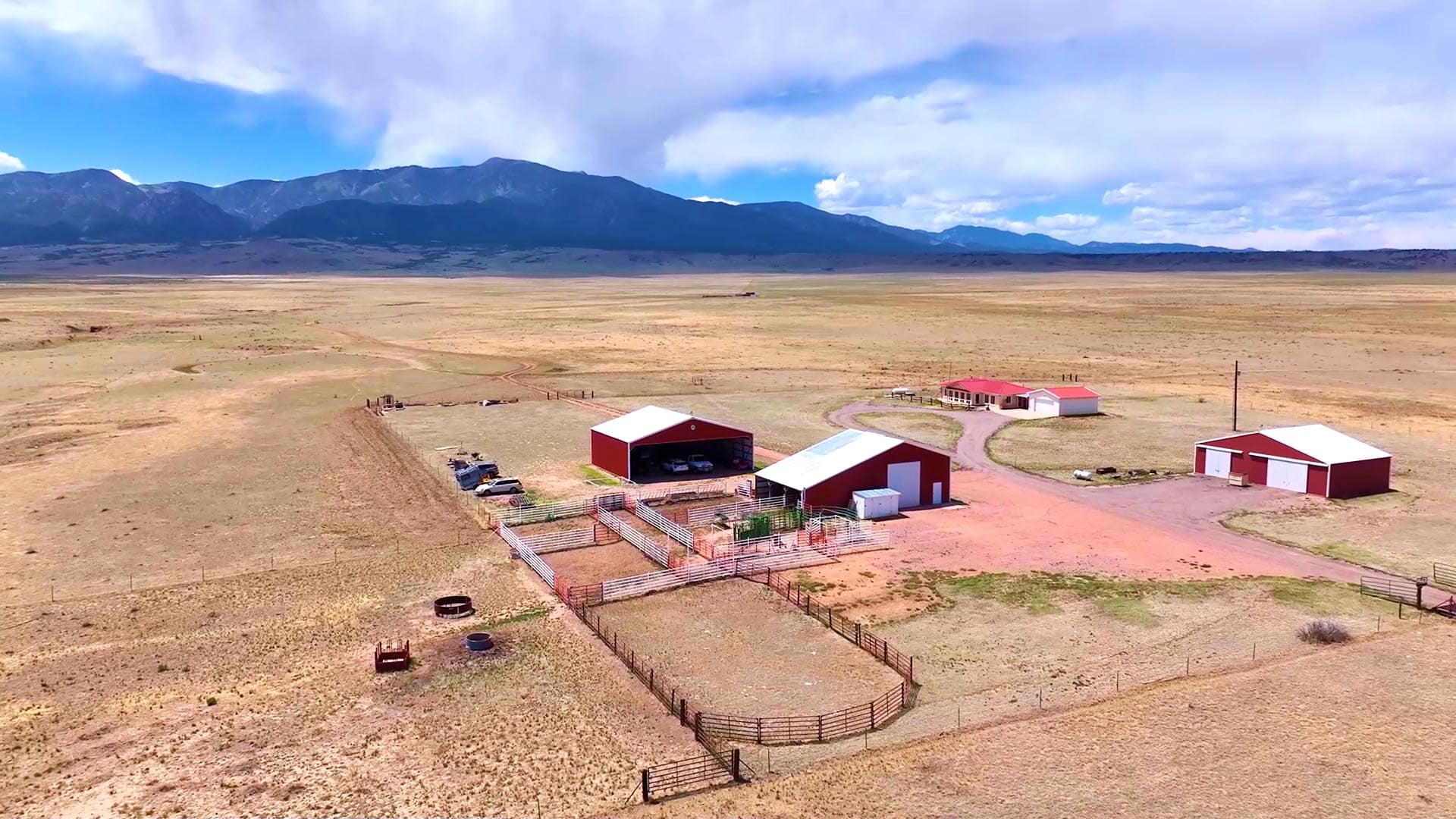 Aerial view of a small ranch with red barns, fences, and dirt roads in a dry, wide valley with distant mountains on the horizon.