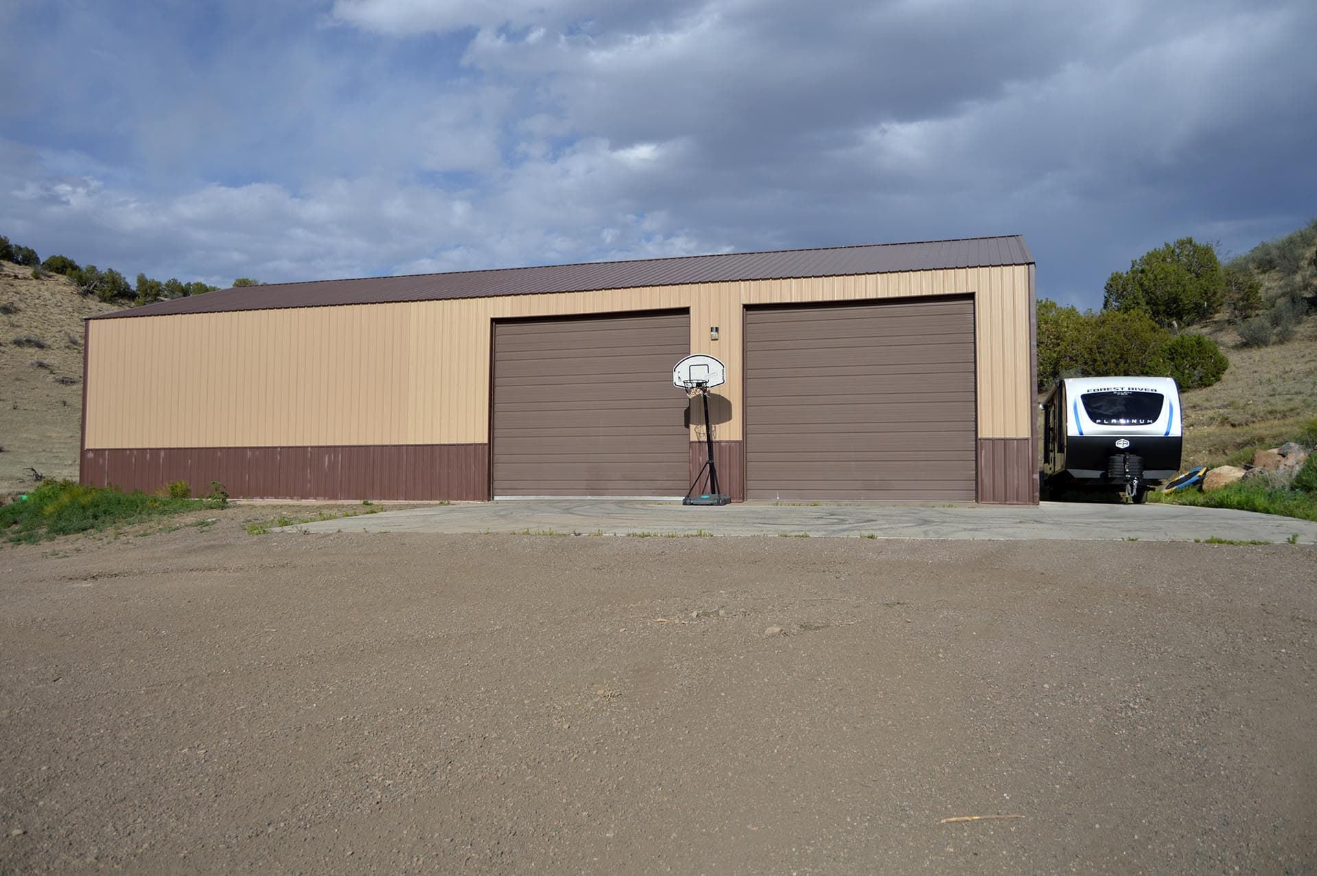 Metal beige storage building with two brown garage doors in a rural lot; a portable basketball hoop stands between doors; a trailer sits to the right under trees.