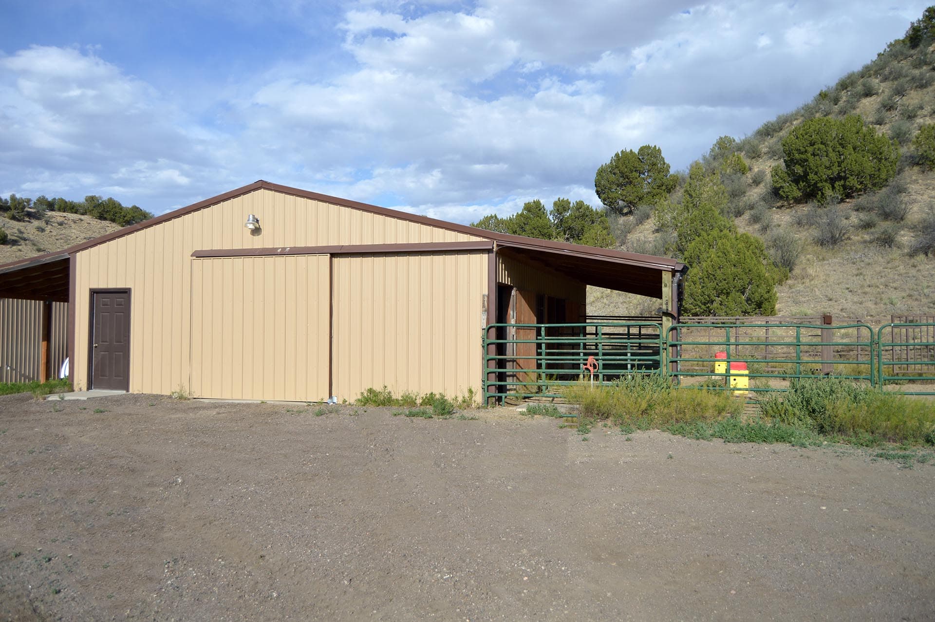 Beige metal-sided building with brown gable roof and a dark door, set in a dirt lot with hills and trees in the background