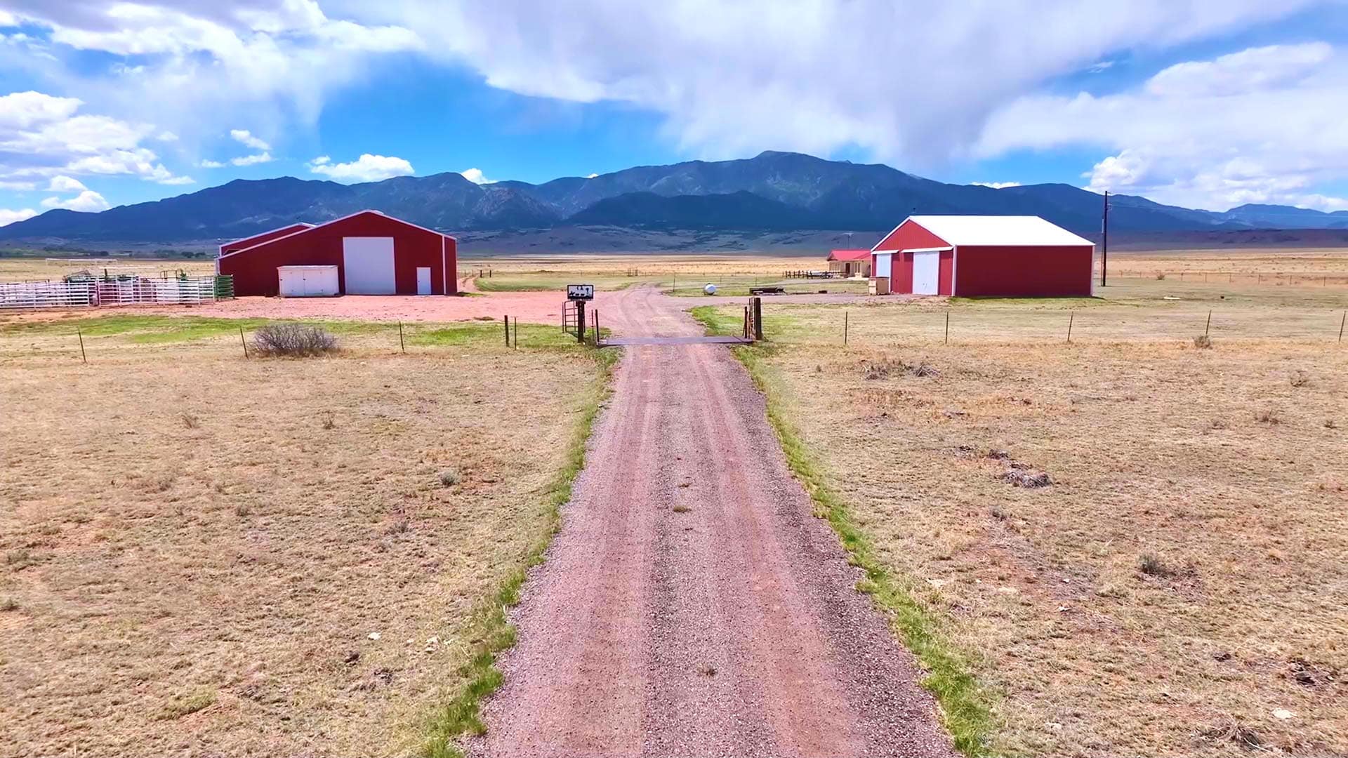 Wide rural farm scene with red barns, gravel road, and distant mountains under a partly cloudy sky