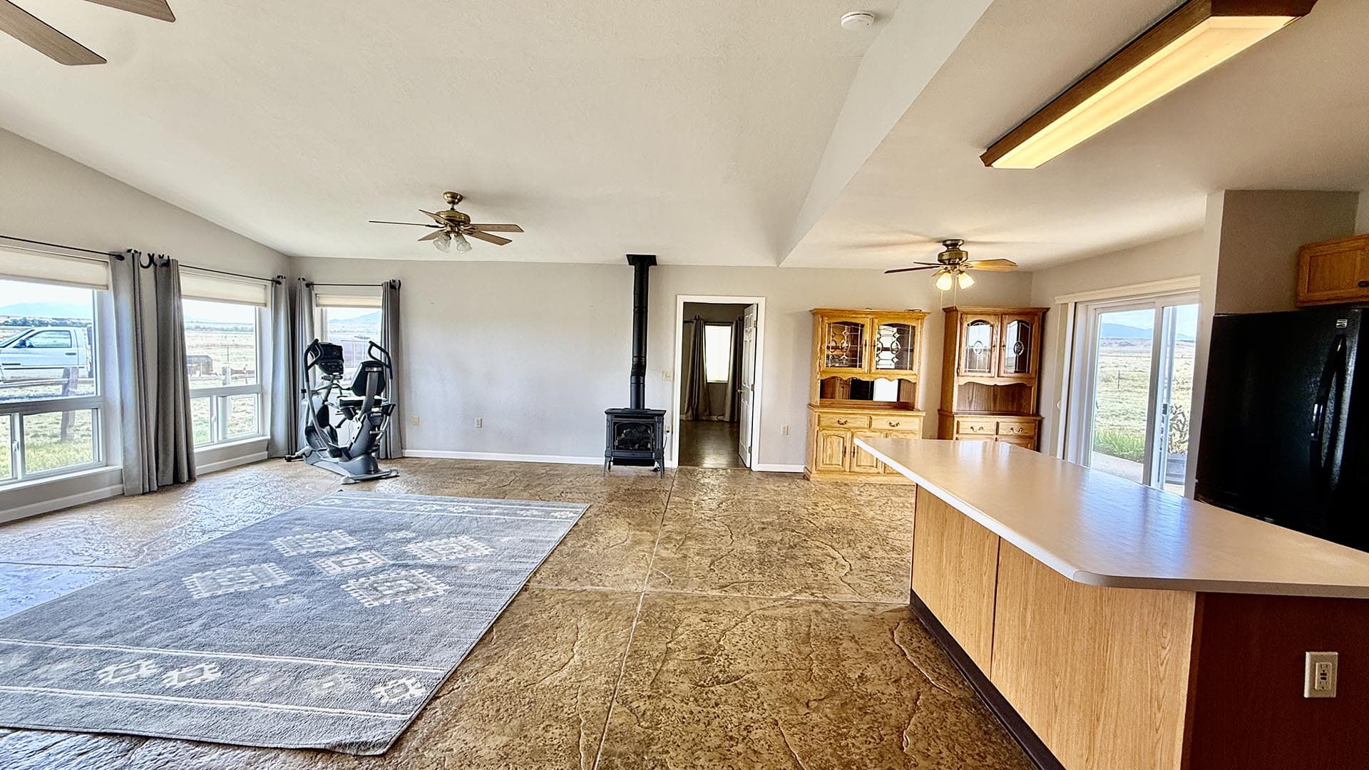 Open living area with large windows, an exercise bike, and a wood stove; tiled floor and a kitchen island in the foreground.