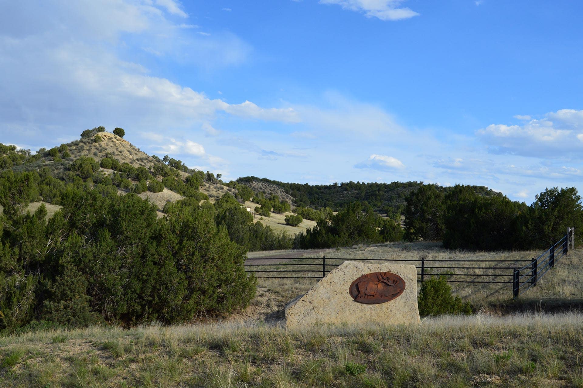 Wide landscape with a grassy foreground, a rocky, tree-covered hill on the left, and a blue sky with clouds; a stone with a circular metal sculpture sits near a fence in the foreground