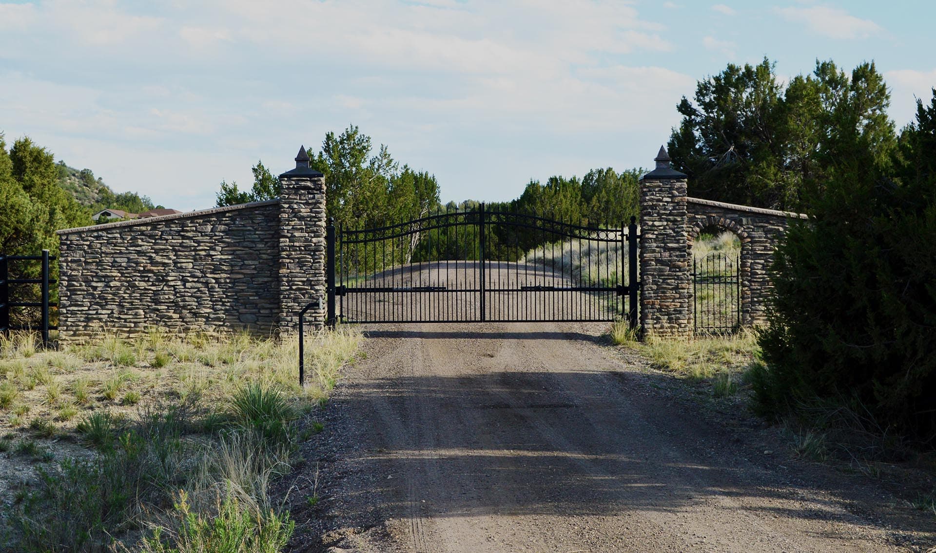 Stone entrance gate with iron gates across a gravel driveway, flanked by stone pillars and desert shrubs.
