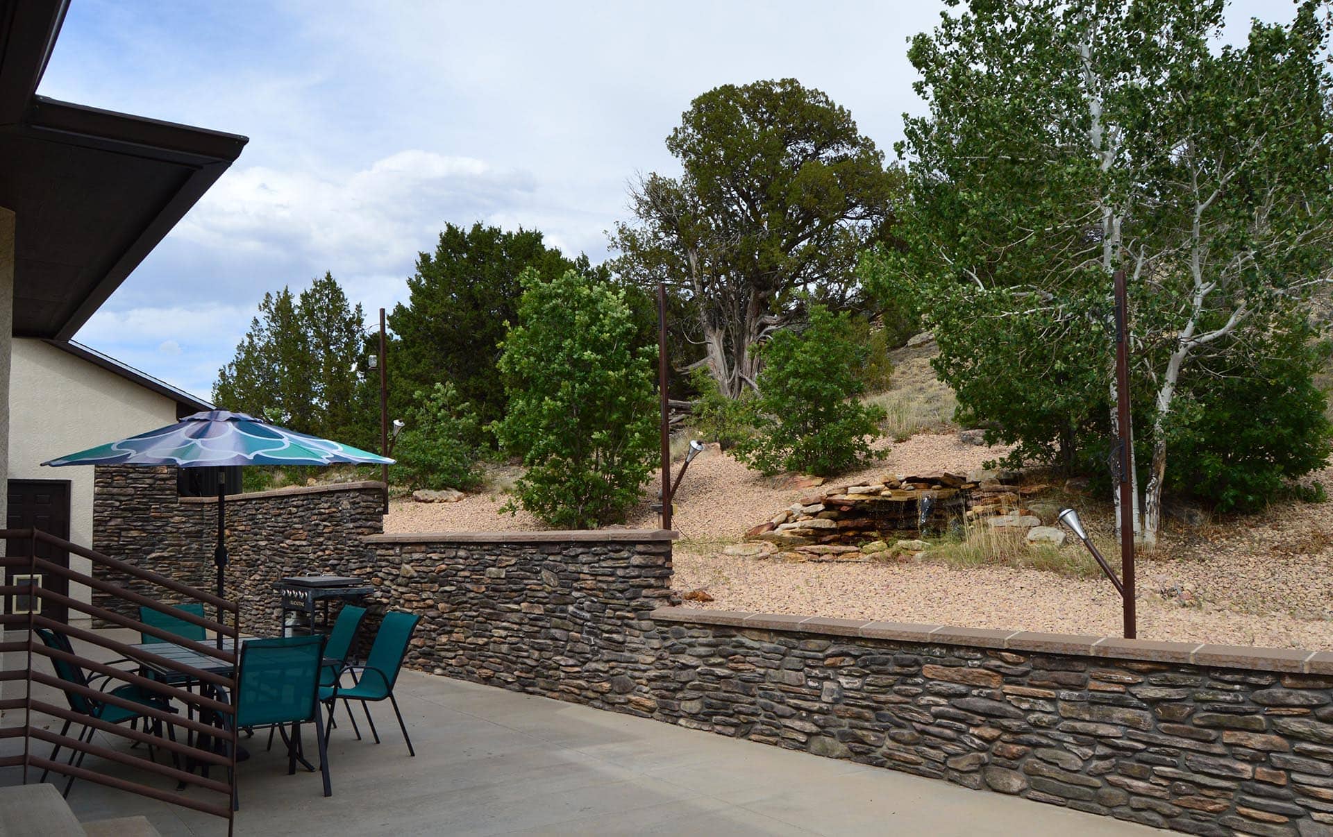 Outdoor seating area next to a stone wall with teal chairs and a blue patterned umbrella, overlooking a rocky hillside with trees.