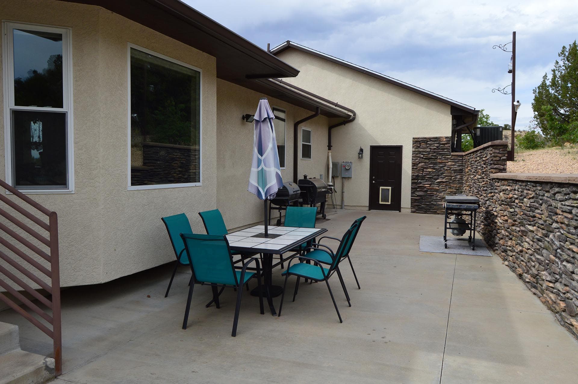 Backyard patio with a dining table, teal chairs, and a folded umbrella next to a beige house wall.