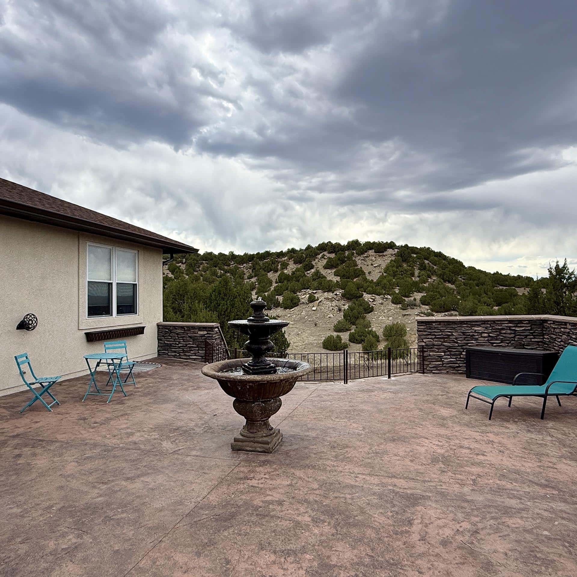 Patio with a central fountain, turquoise chairs, and a beige house wall, overlooking a rocky, tree-covered hillside under a cloudy sky.