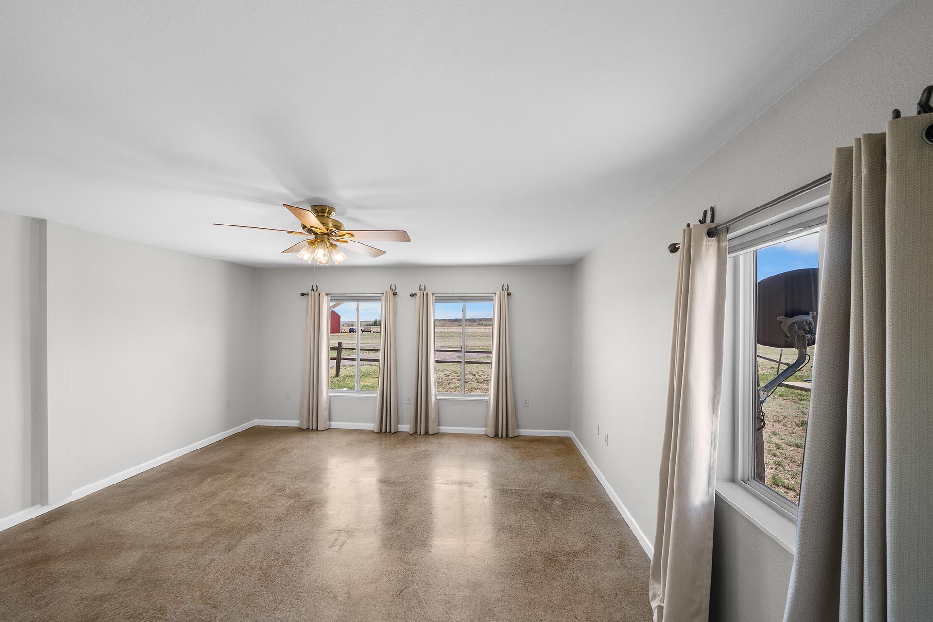 Empty living room with beige carpet, light gray walls, a ceiling fan, and windows with beige curtains looking out onto a rural landscape.
