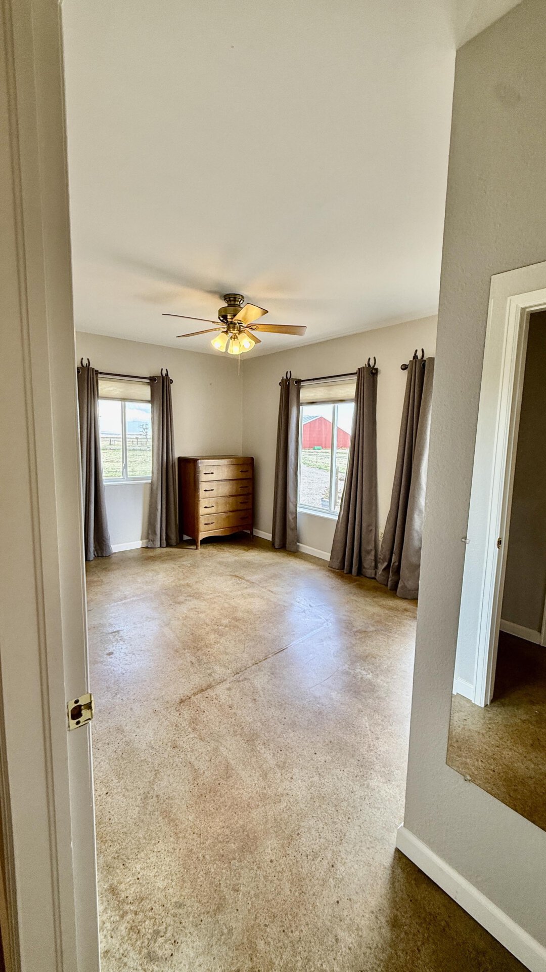 Bedroom with beige carpet, two windows with gray curtains, and a wooden dresser under the corner window; ceiling fan above.