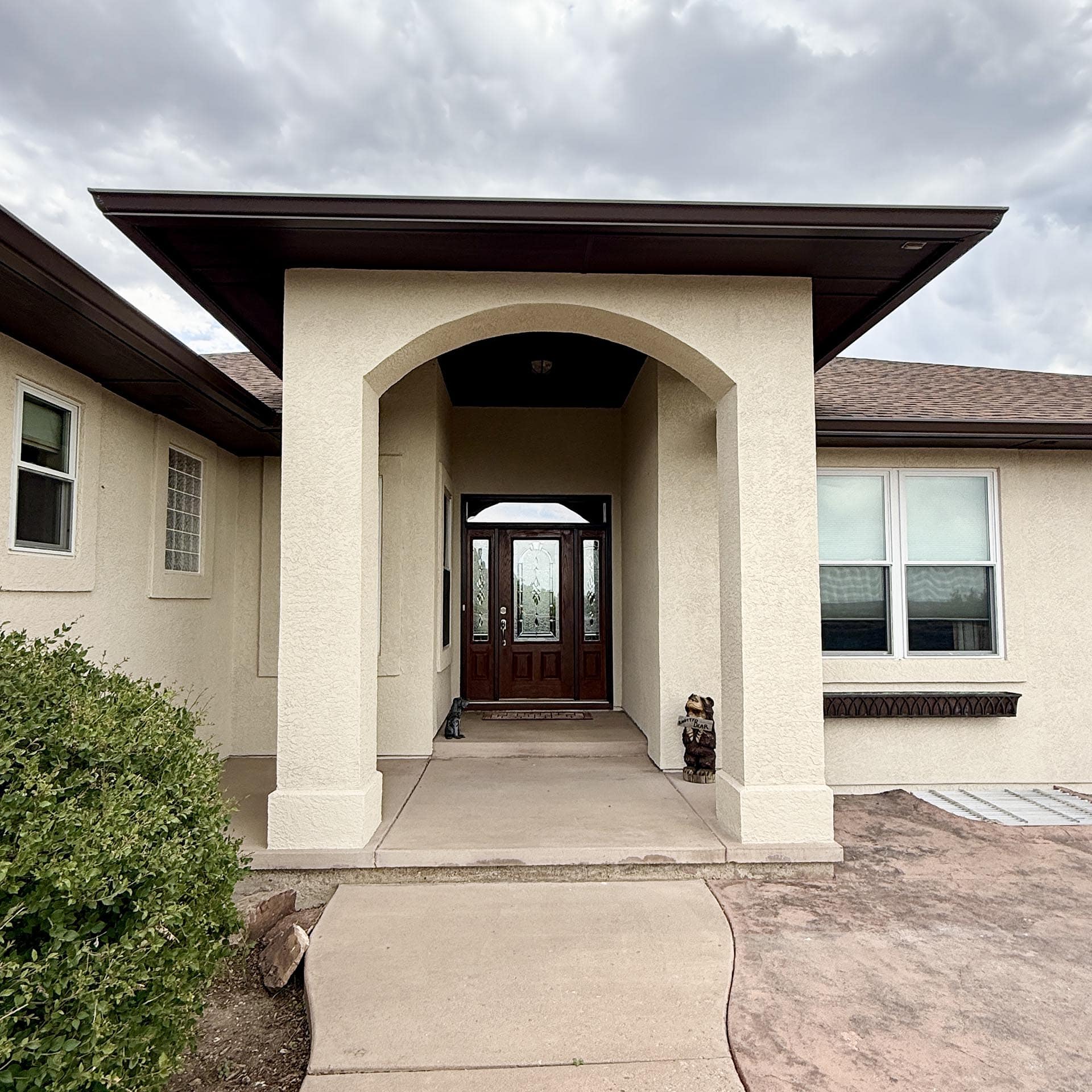 Residential entrance with beige stucco walls, arched entryway, and dark wooden front door under a covered porch.