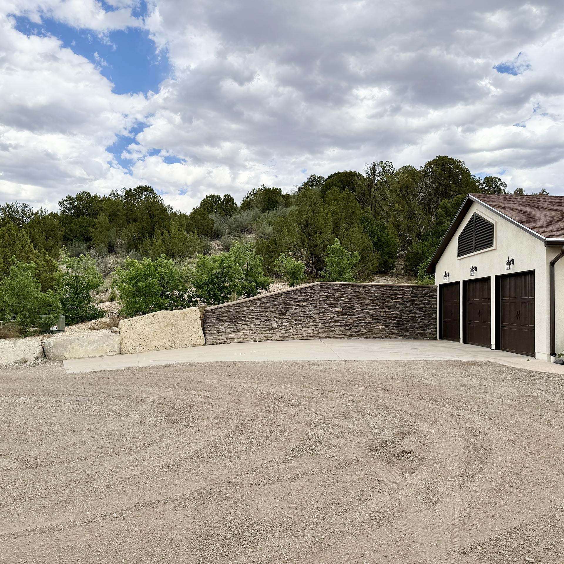 Beige house with dark garage doors beside a stone retaining wall and gravel driveway, hillside and trees behind under a cloudy sky.