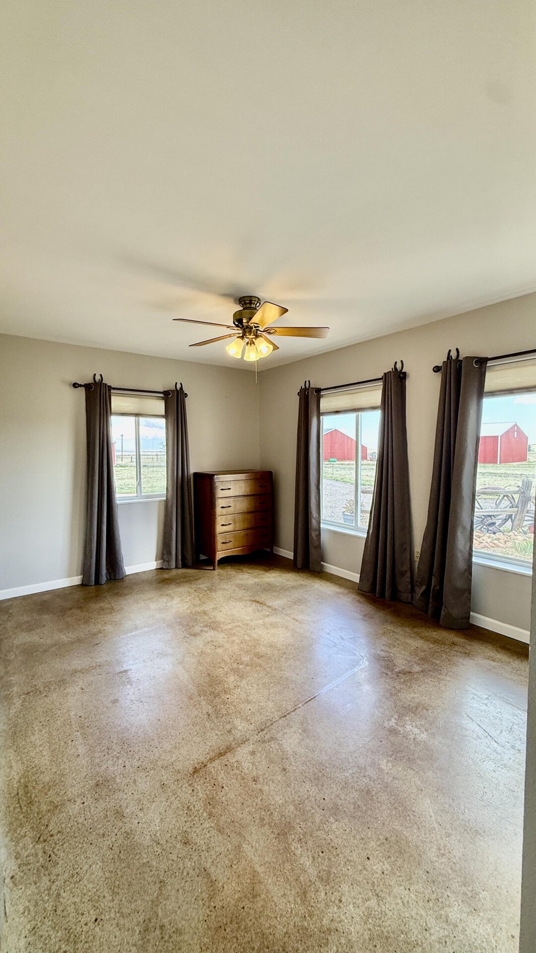 A bright empty bedroom with a ceiling fan, a wooden dresser, and three windows with dark curtains showing a rural yard and red barns outside; neutral walls and brown carpet.