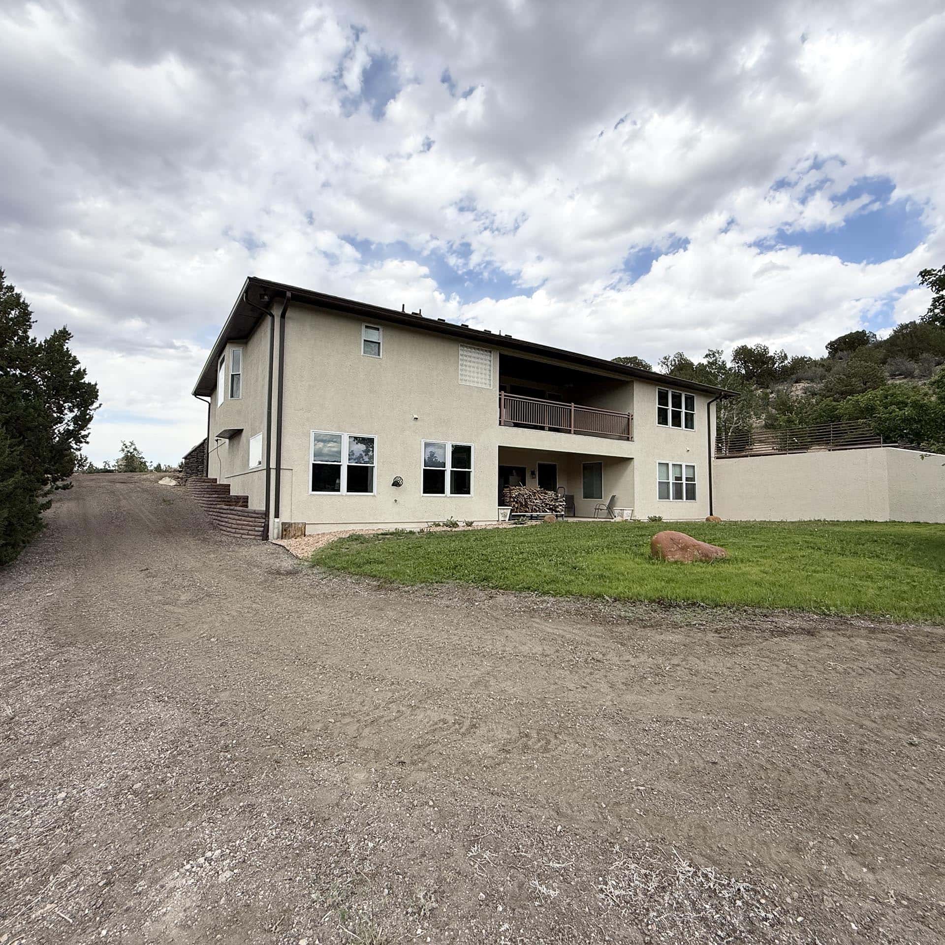 Two-story beige house with a brown balcony on a hillside, gravel driveway in the foreground and a pile of firewood outside.